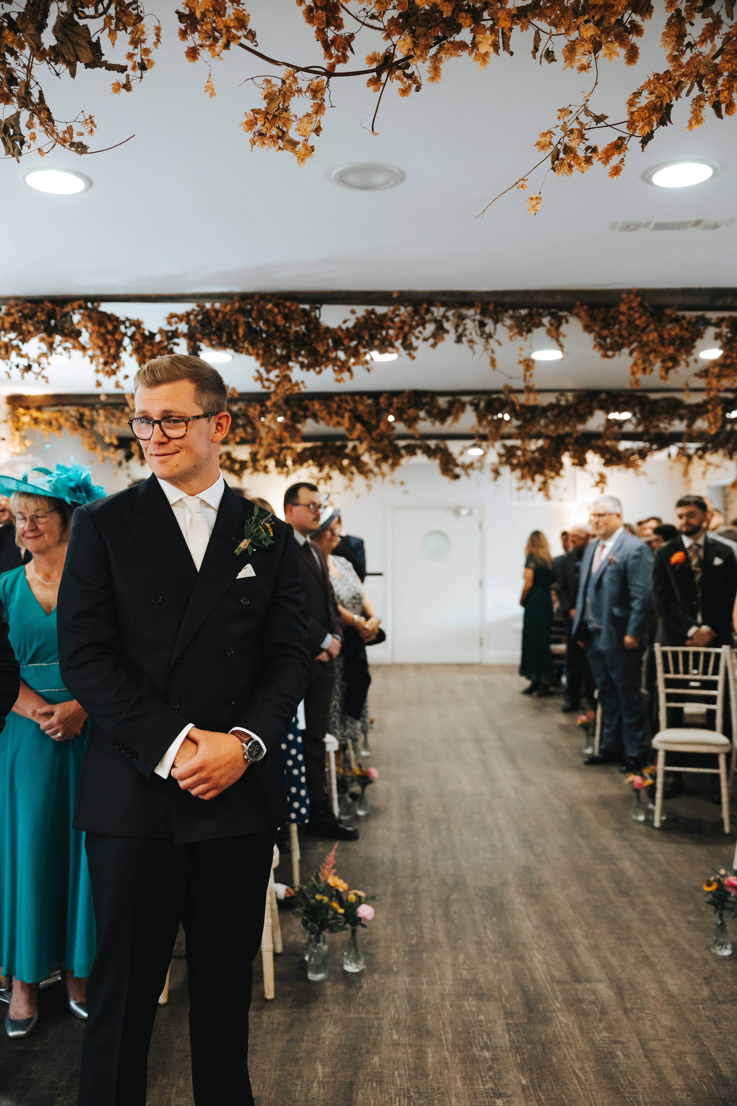 Young man in a tuxedo standing at a wedding ceremony, surrounded by seated guests in formal attire, with autumn-themed decorations hanging from the ceiling.