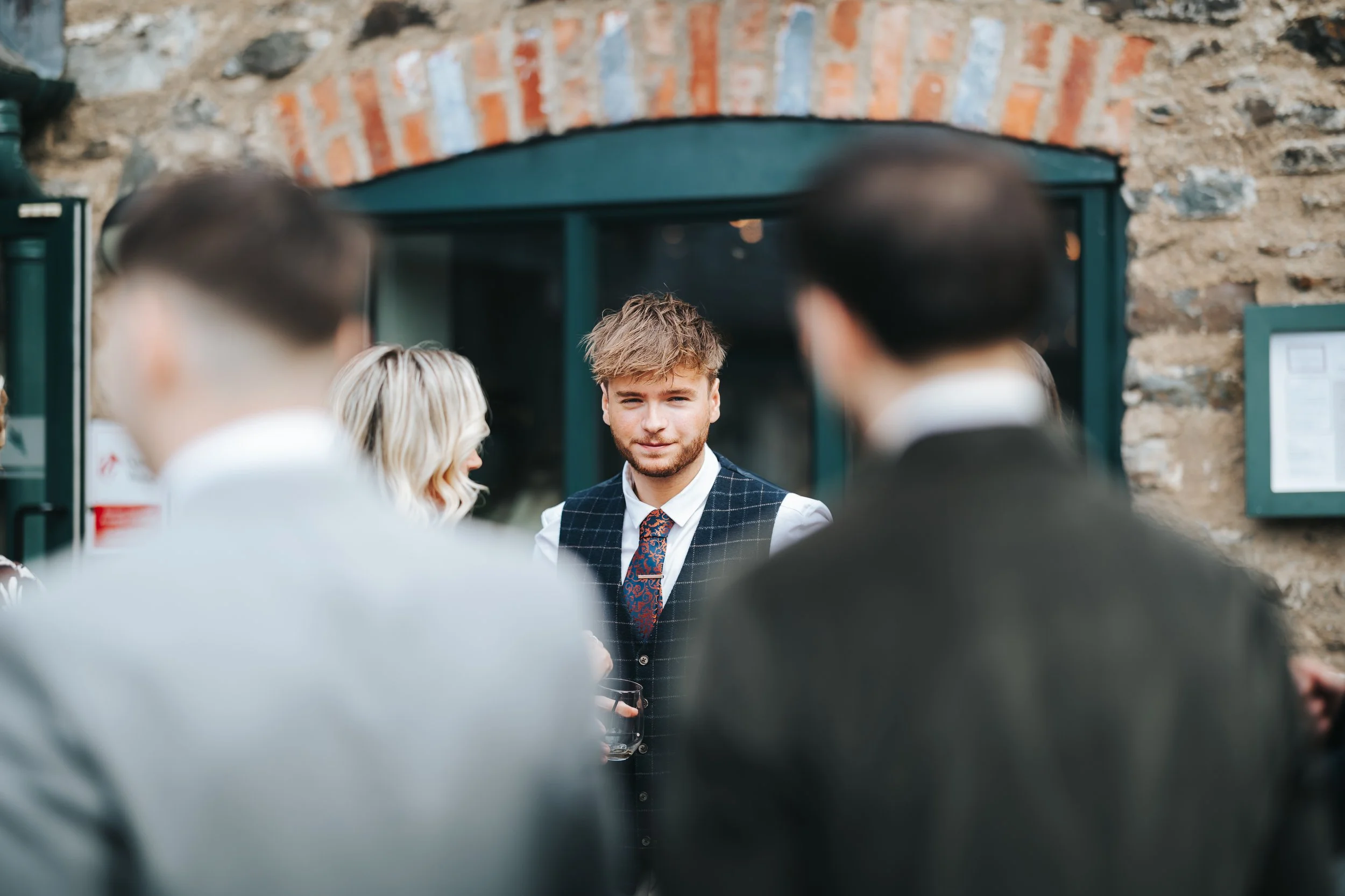 A young man with light brown hair and a beard, wearing a dark vest, white shirt, and a colorful tie, standing outdoors in front of a brick building with a green door, surrounded by people dressed in formal attire, at a social gathering or wedding rec