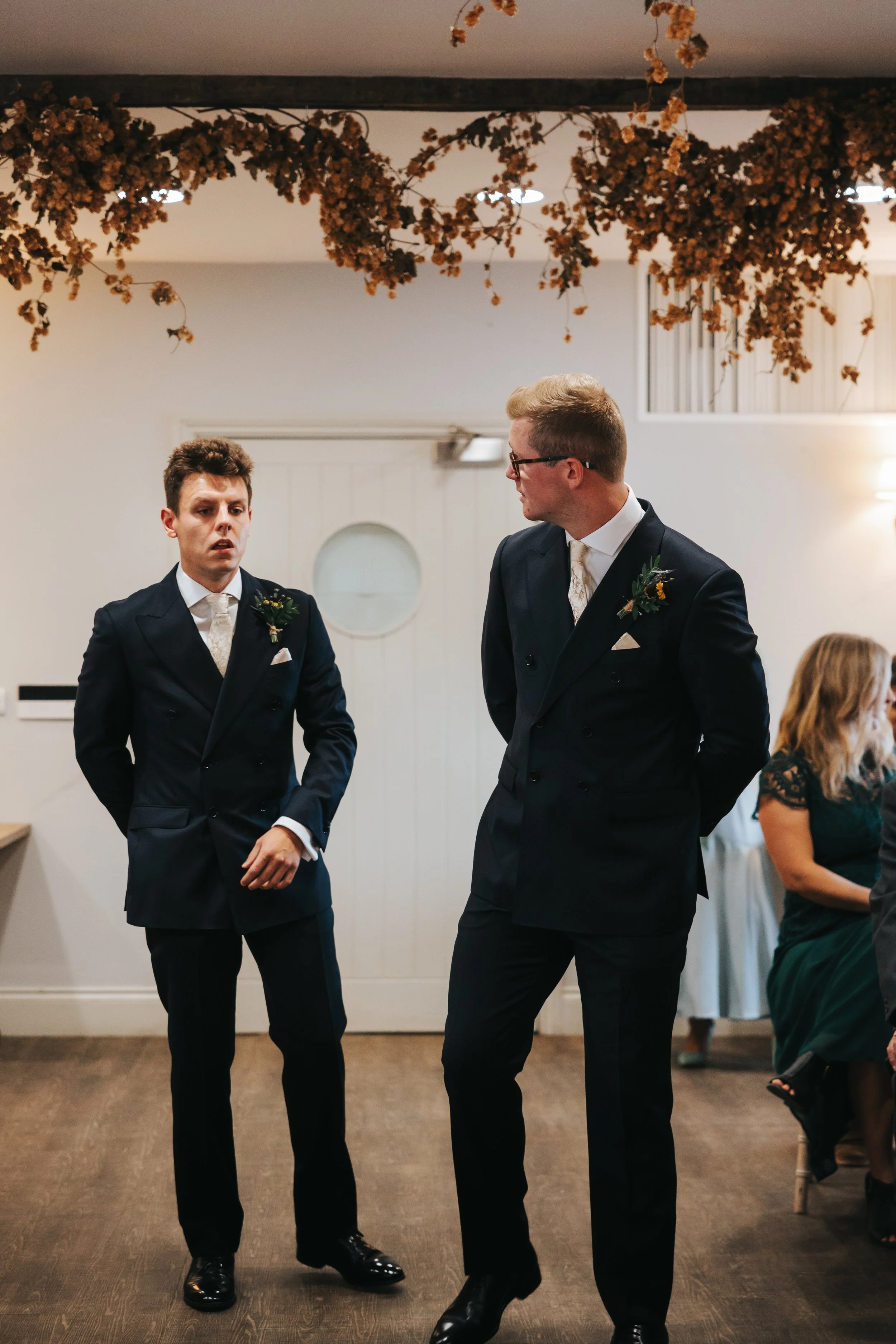 Two men in dark suits and white shirts standing inside a room decorated with hanging dried flowers, engaged in conversation or dancing, with people seated at tables in the background.
