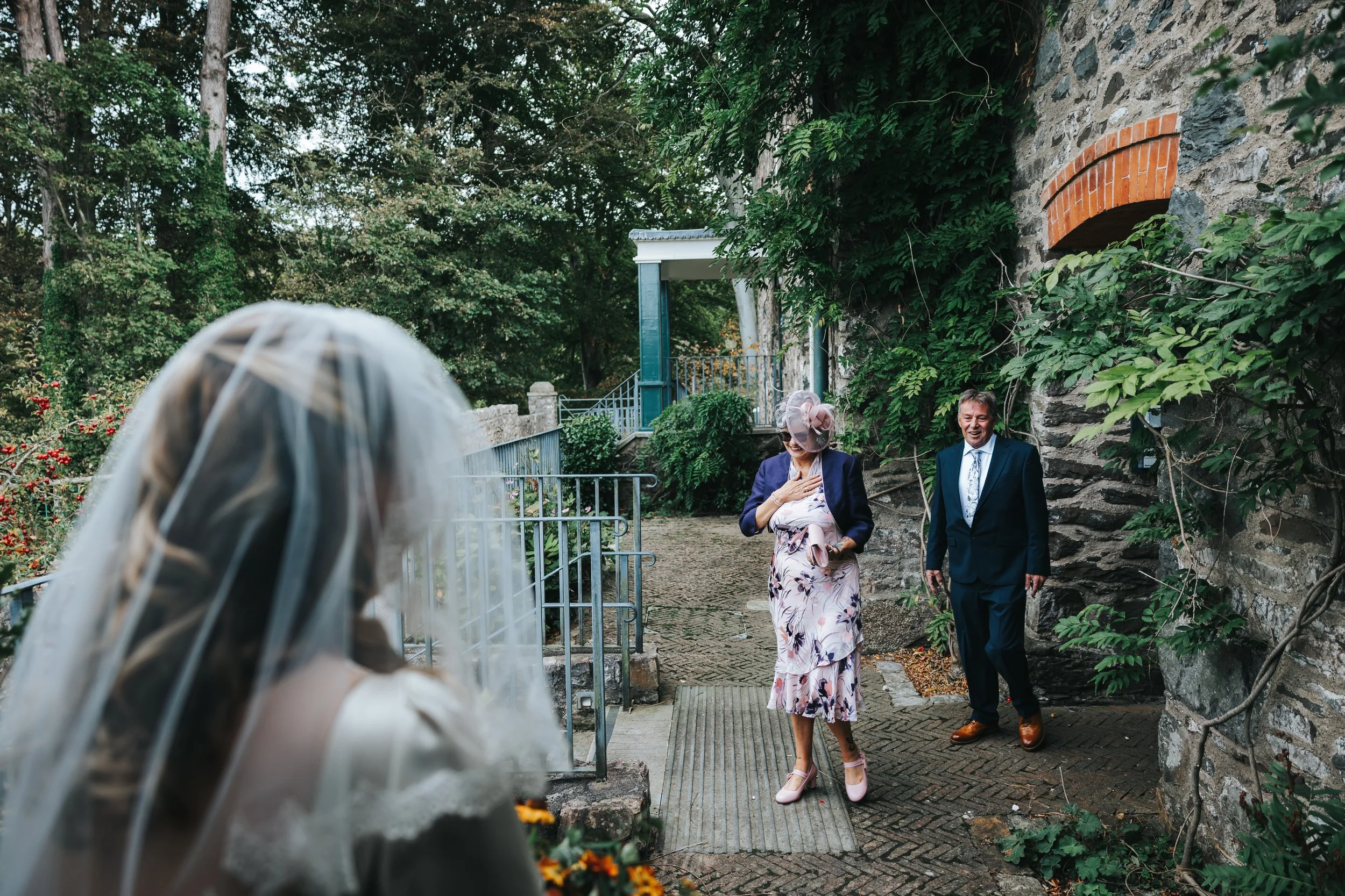 A bride with a veil and floral bouquet faces away from the camera, observing two guests, a woman in a floral dress and hat and a man in a suit, walking and smiling on a garden pathway with greenery and stone walls.
