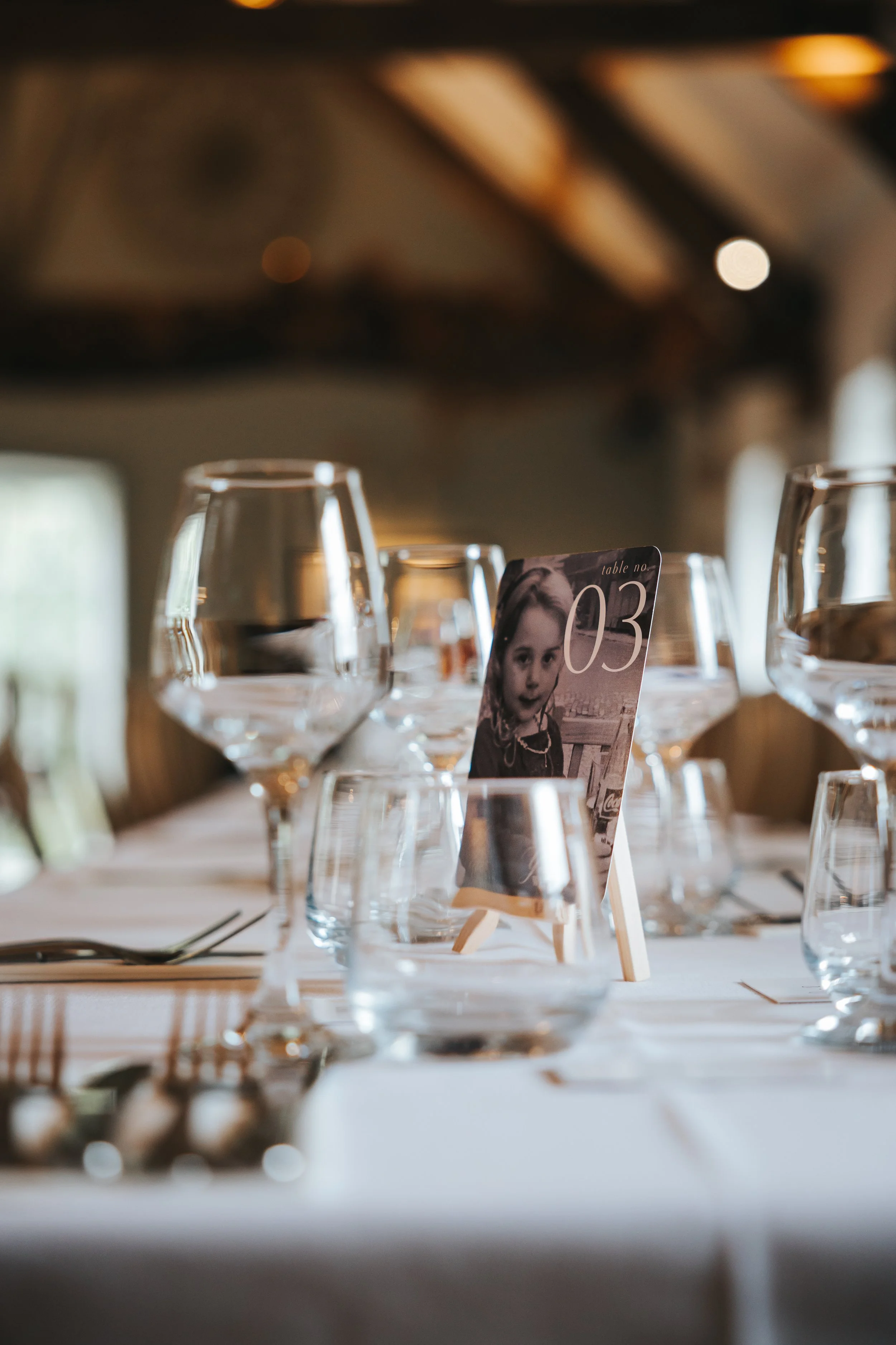 Elegant dining table set with clear wine glasses, a table number card showing a child's black-and-white photo with a surprised expression, and neatly arranged cutlery in a warmly lit venue.