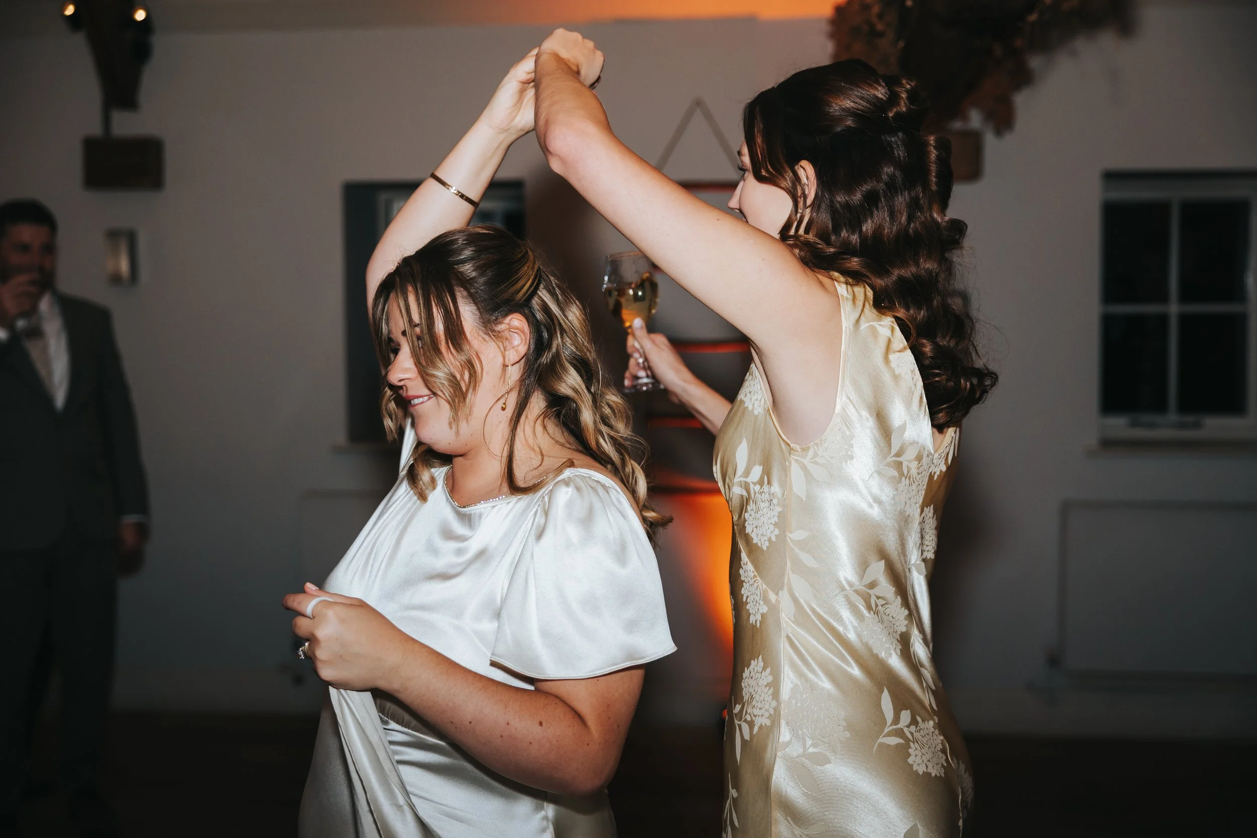 Two women dancing at a celebration, with one woman holding a wine glass, wearing elegant dresses, in a decorated indoor setting, with a man in a suit in the background.