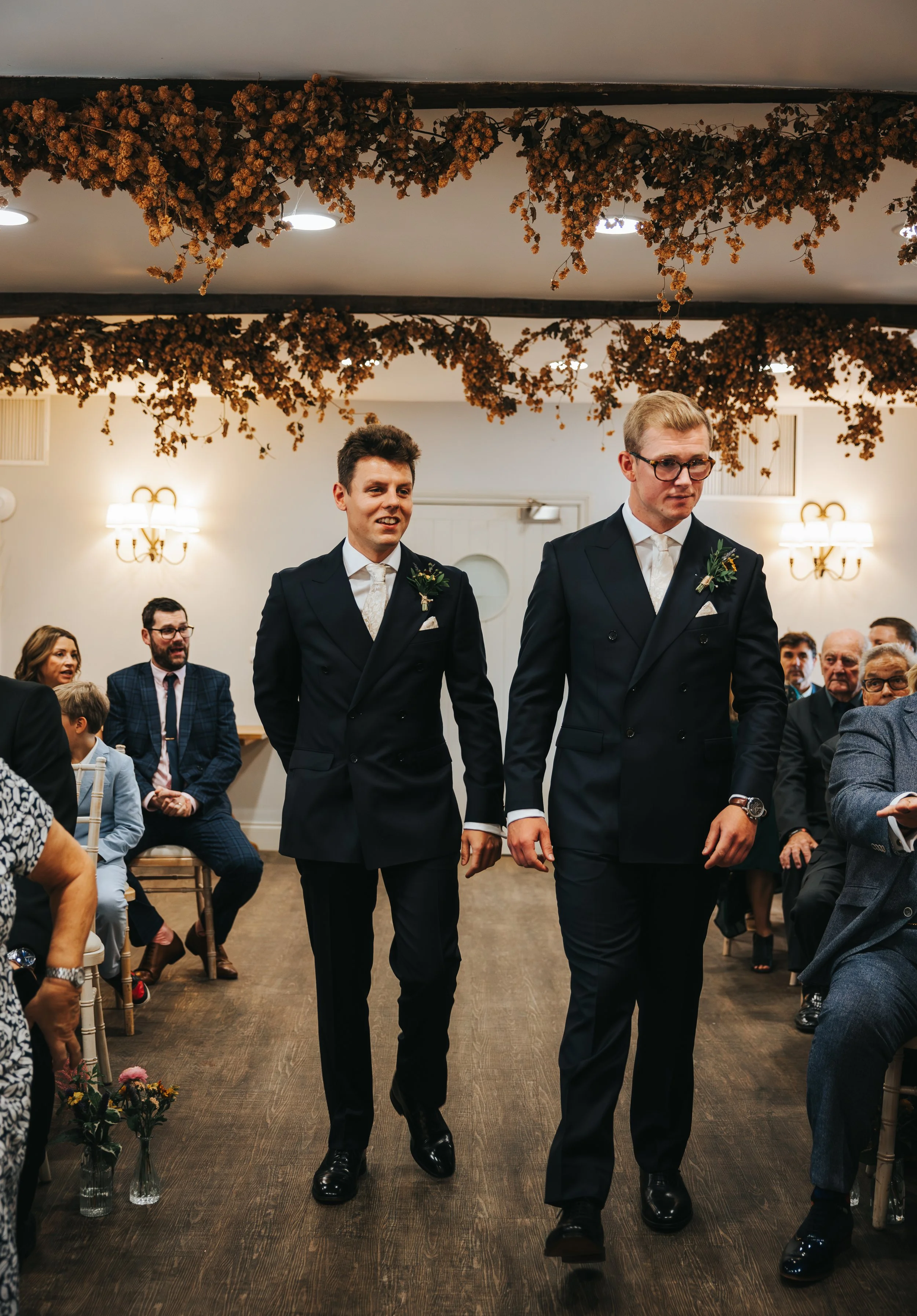 Two men in suits walking down an aisle during a wedding, with seated guests watching.