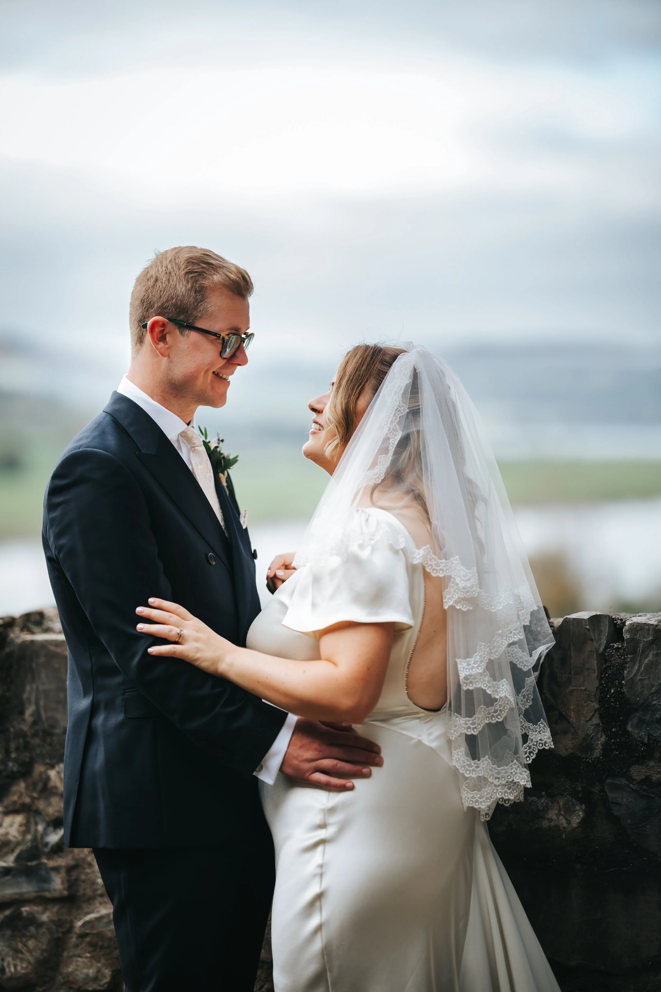 A bride and groom on their wedding day, smiling at each other outdoors, with a rocky wall and a body of water in the background.
