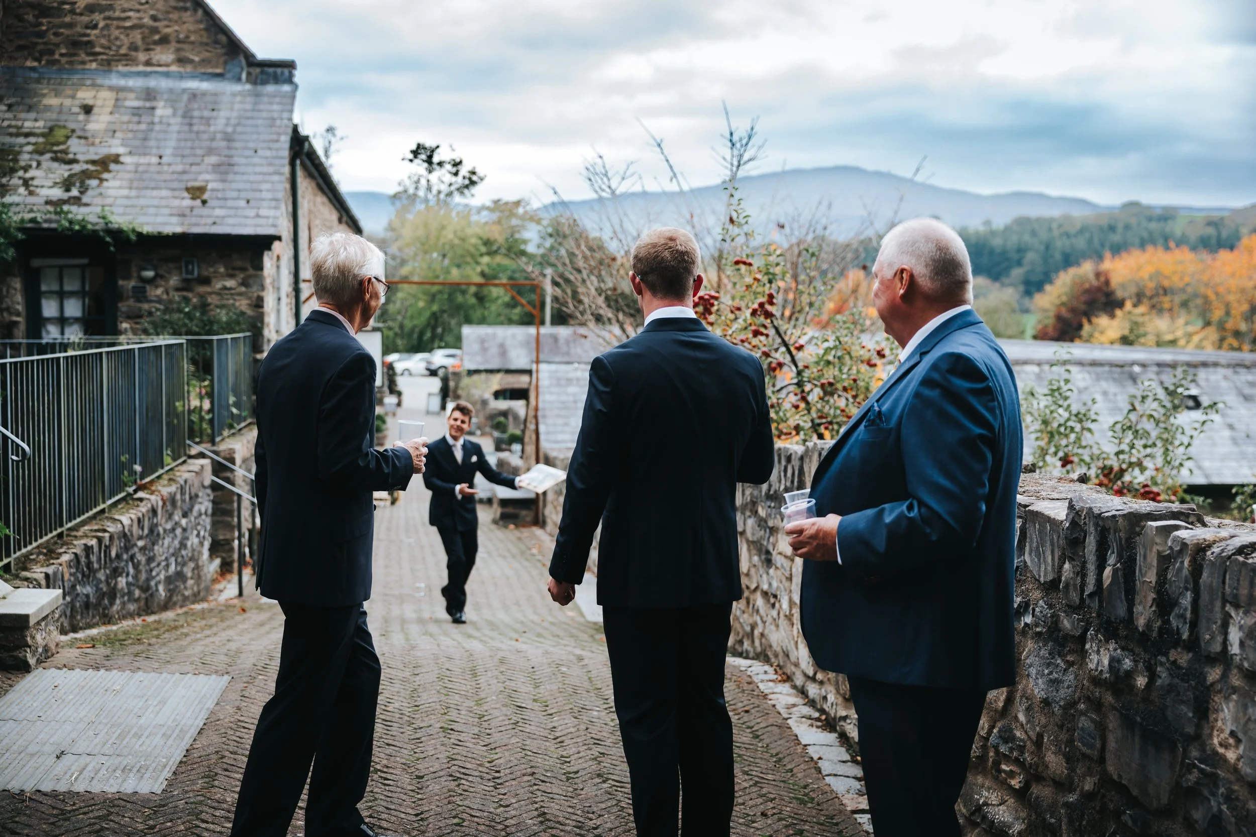 Four men in suits standing on a cobblestone path outdoors, with a stone wall, trees, and mountains in the background. One man is running towards the group holding a magazine or booklet, and the others are talking or holding drinks.