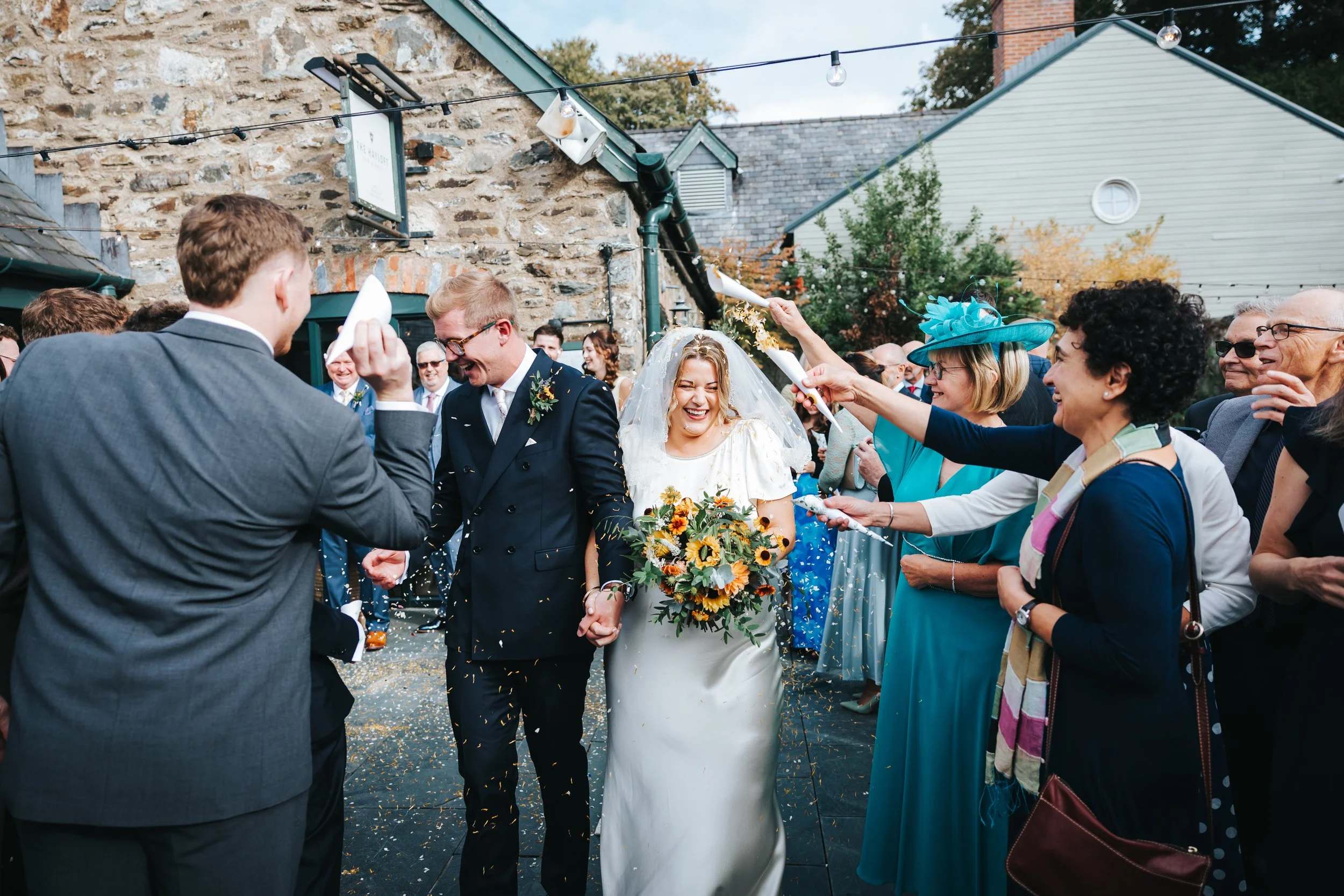 Bride and groom walking hand in hand through a shower of confetti, smiling, with guests throwing confetti and celebrating outdoors.