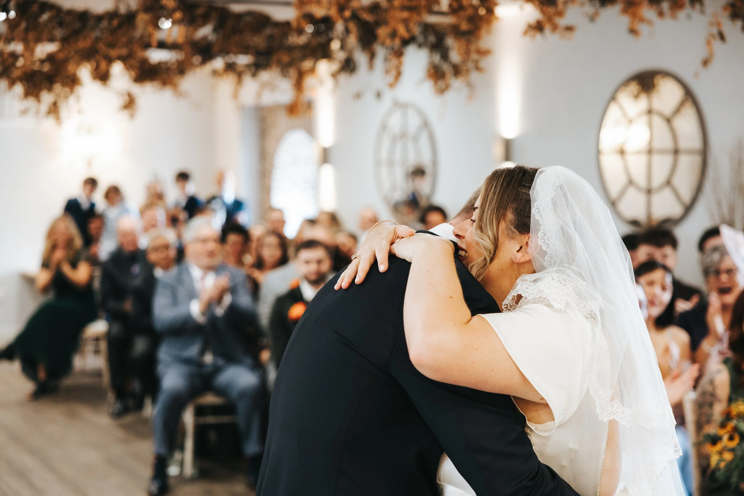 A bride and groom embrace tightly during a wedding ceremony, with guests clapping and smiling in the background inside a decorated venue.