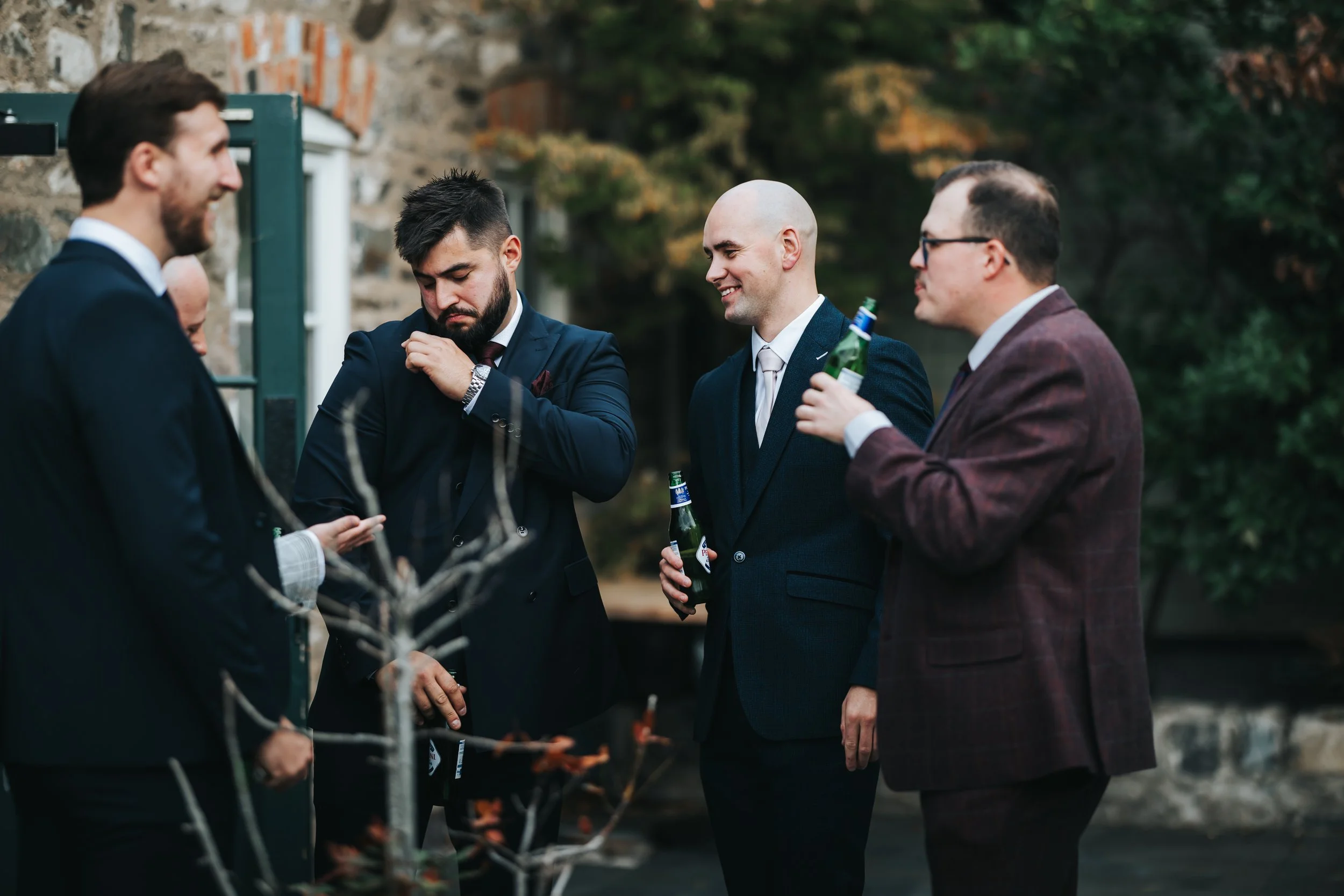 Group of five men in suits holding beer bottles, engaged in conversation outdoors near a stone building and trees.