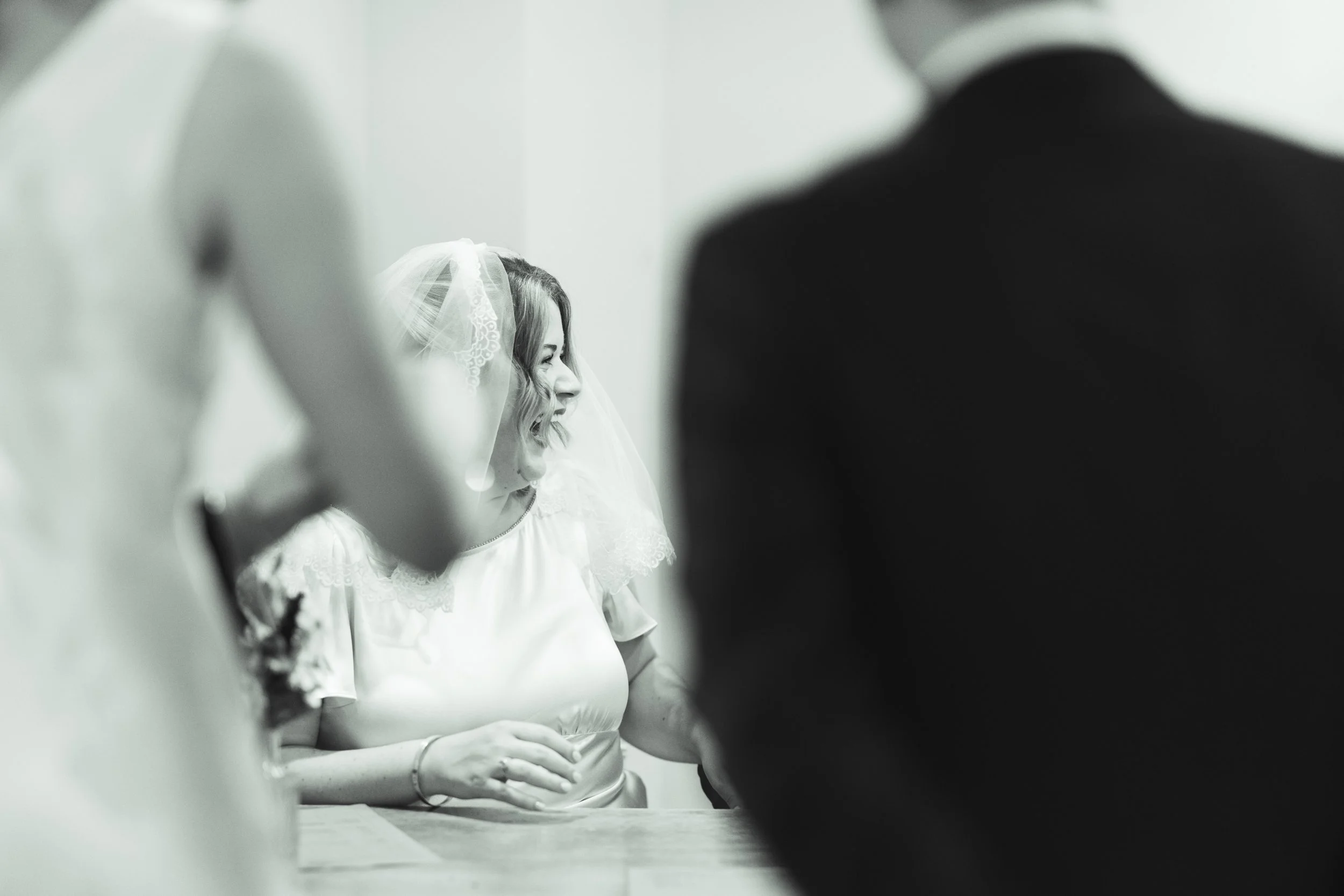 A black and white photo of a bride and an officiant during a wedding ceremony, with the bride smiling and wearing a veil, while the groom is partially visible in the foreground.