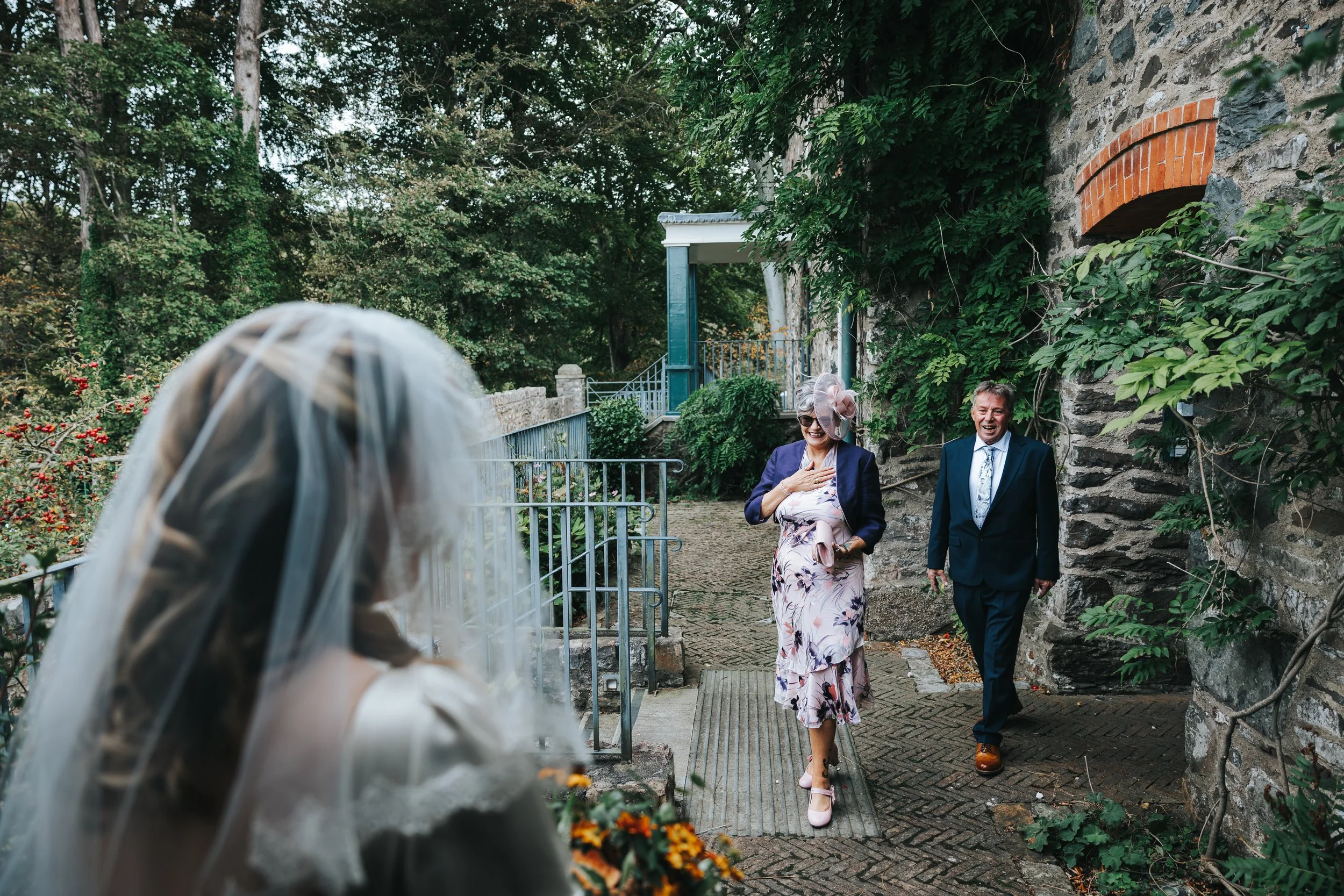 A woman dressed as a bride with a veil, holding a bouquet, is facing a couple walking towards her. The couple, a woman in a floral dress and a man in a suit, are smiling and appear surprised to see her. They are outside near a stone wall covered in g