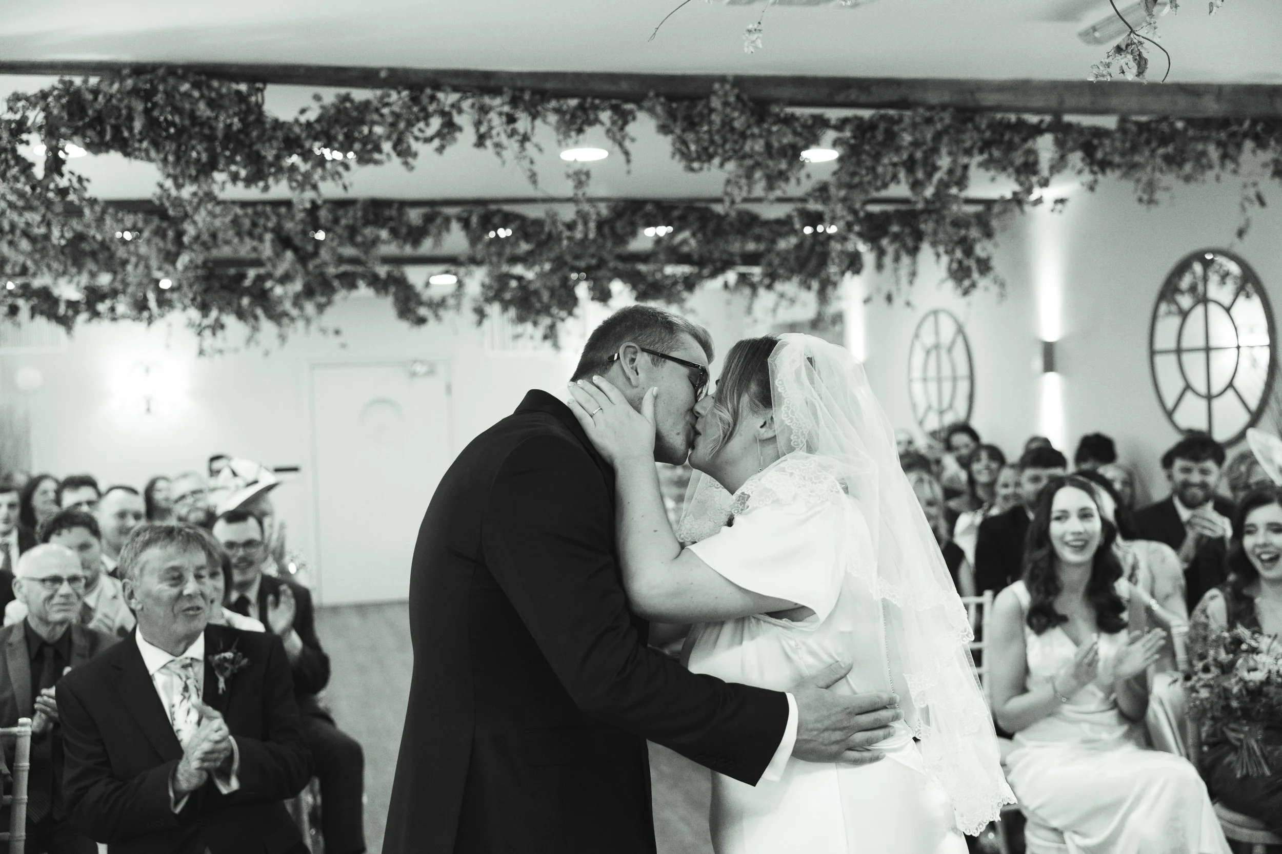 A black-and-white photo of a couple sharing a tender kiss during a wedding ceremony in a decorated indoor venue, with seated guests watching and clapping in the background.