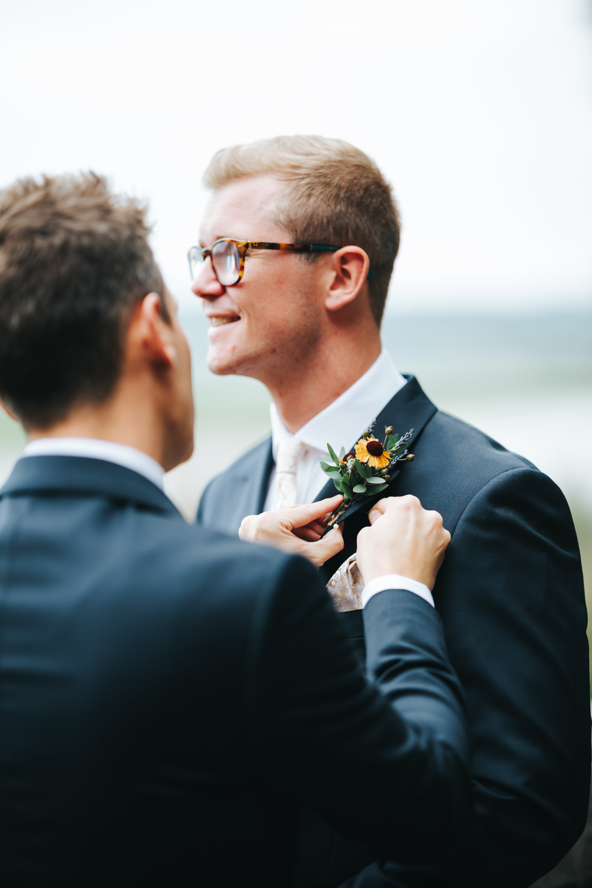 Two men in formal suits sharing an intimate moment, one adjusting the other's boutonniere at a wedding or formal event.