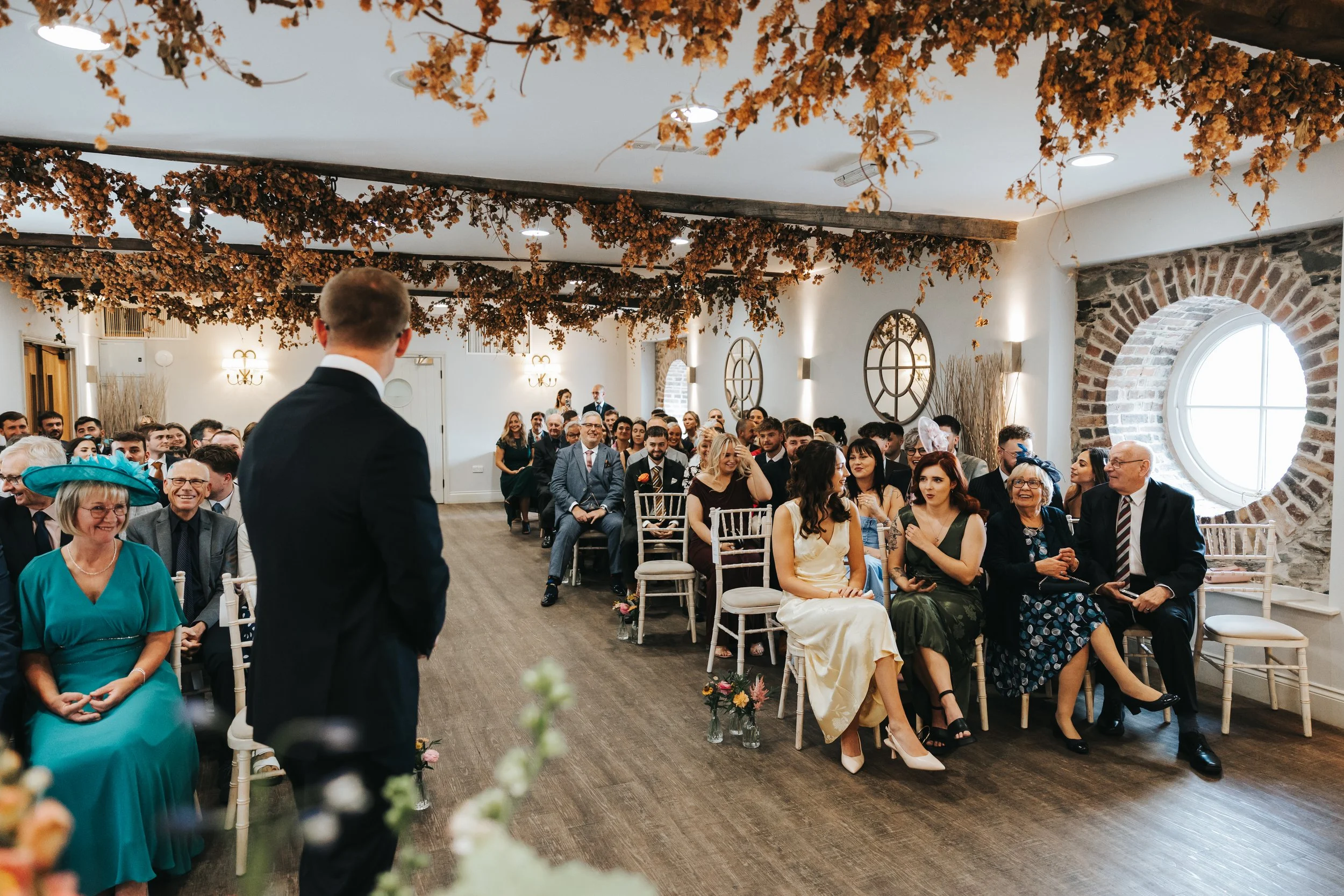 A wedding ceremony with guests seated in chairs, some clapping and smiling, in a decorated venue with hanging flowers and brick walls.