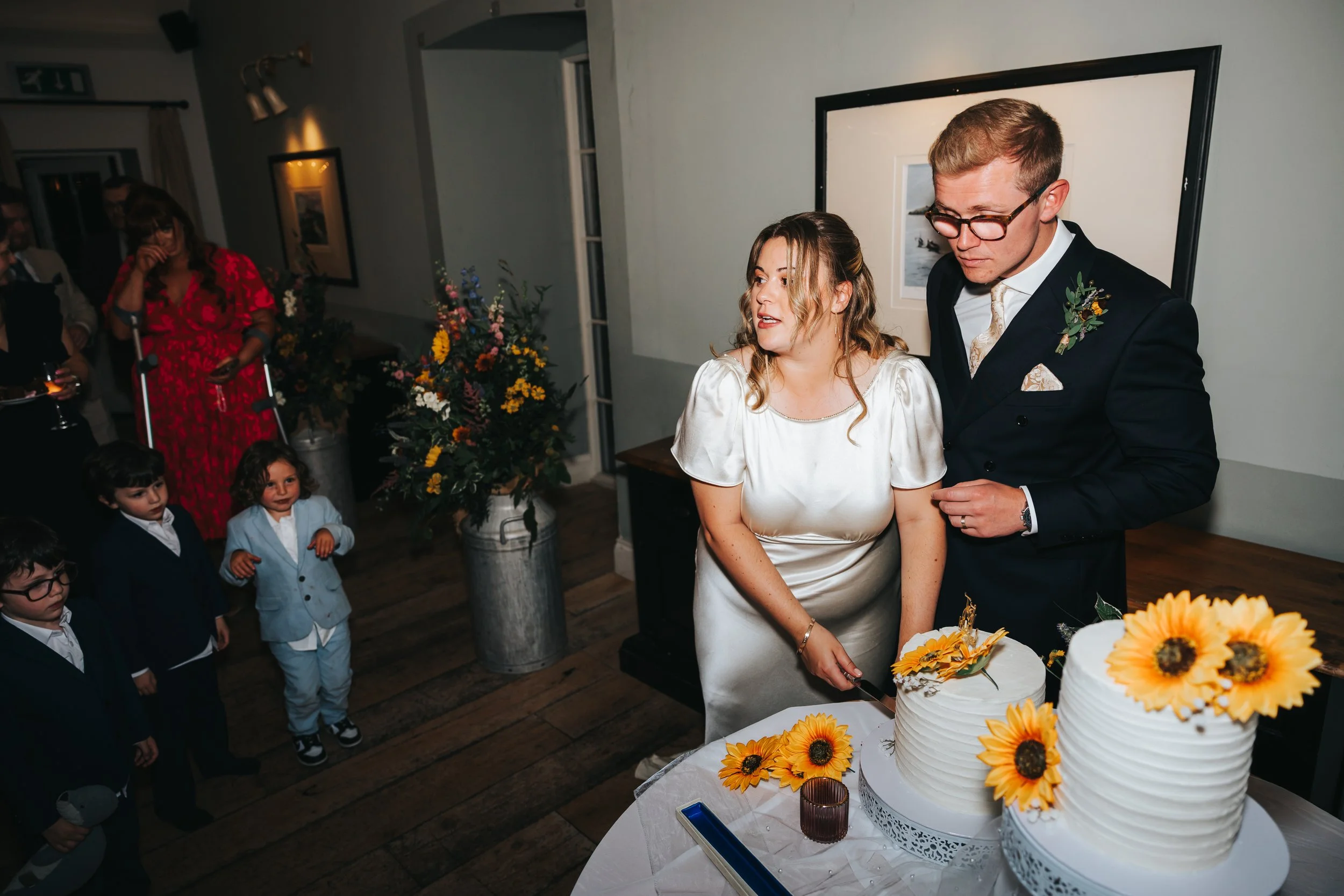 A bride and groom cutting wedding cakes decorated with sunflowers at their wedding reception, surrounded by guests including children and adults.