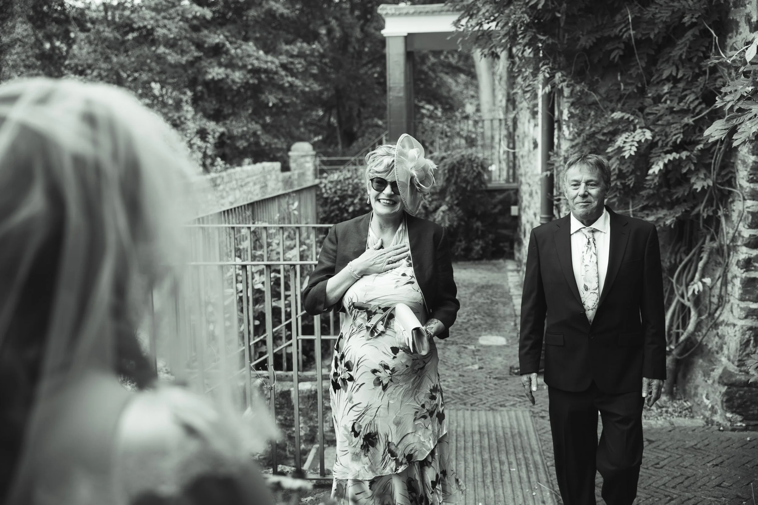 A black and white photo of a woman in a floral dress and a hat with sunglasses smiling and holding her hand to her chest, walking outdoors with a man in a suit and tie, while part of another person's head is visible in the foreground.
