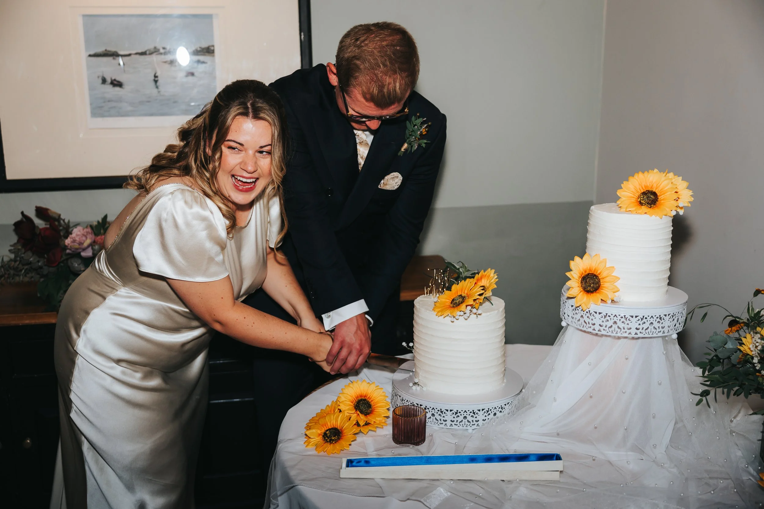 A bride and groom cut a wedding cake together, smiling and holding a knife.