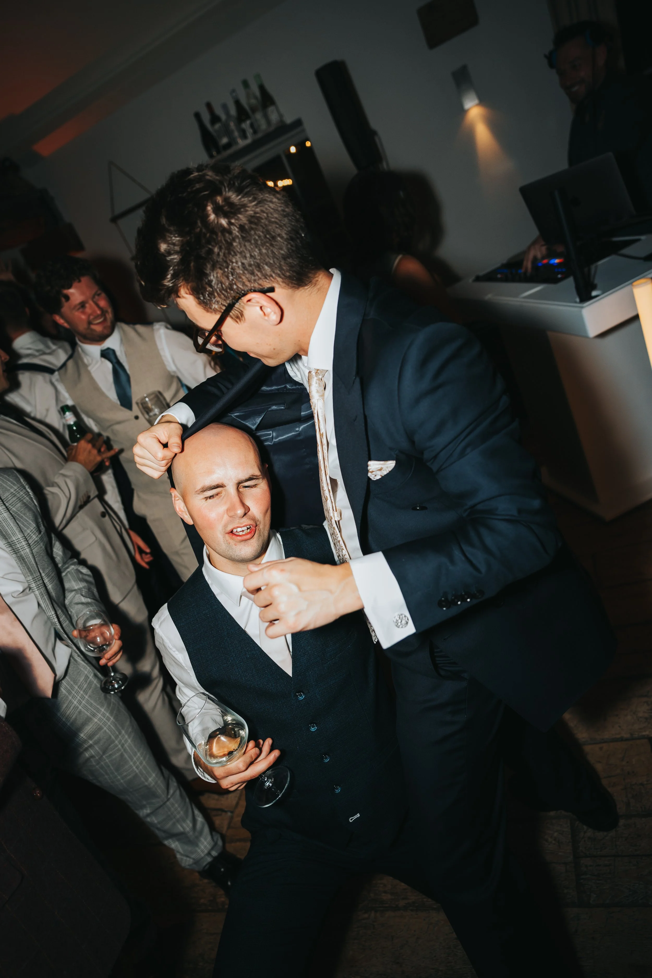 Two men dressed in suits are dancing and having fun at a party with people in the background, some holding drinks.