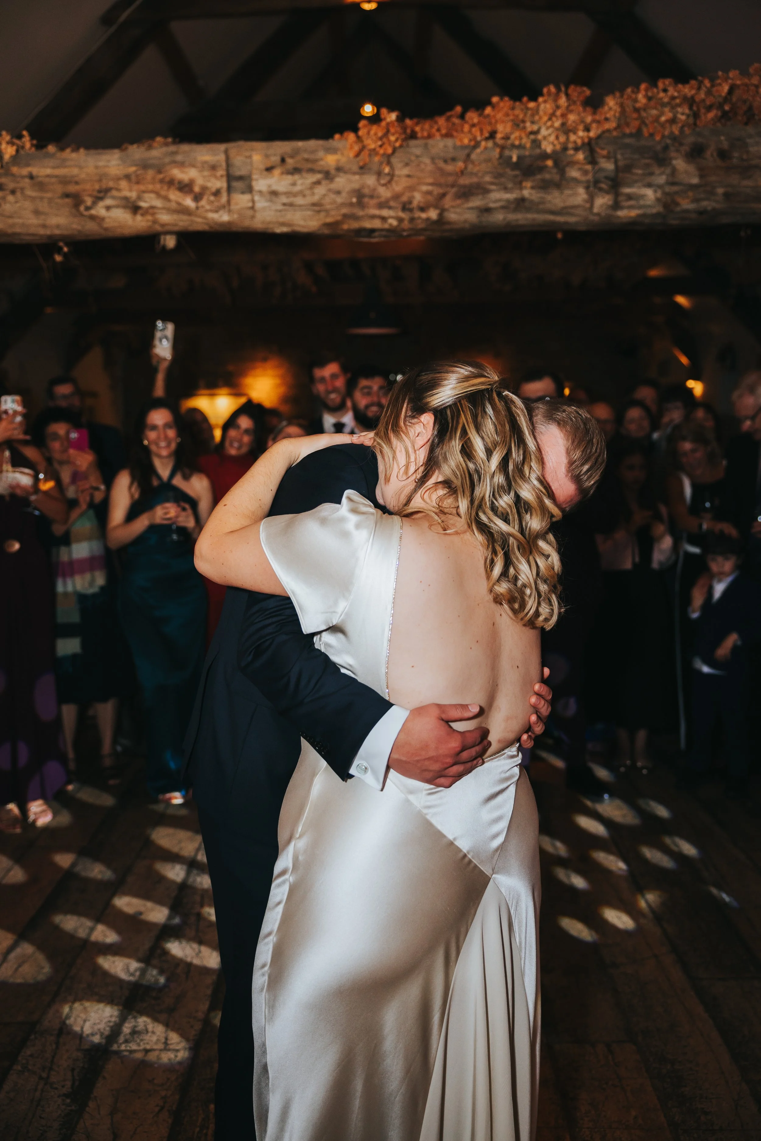 A bride and groom share a dance, embracing each other closely at their wedding reception with guests watching and smiling in the background.