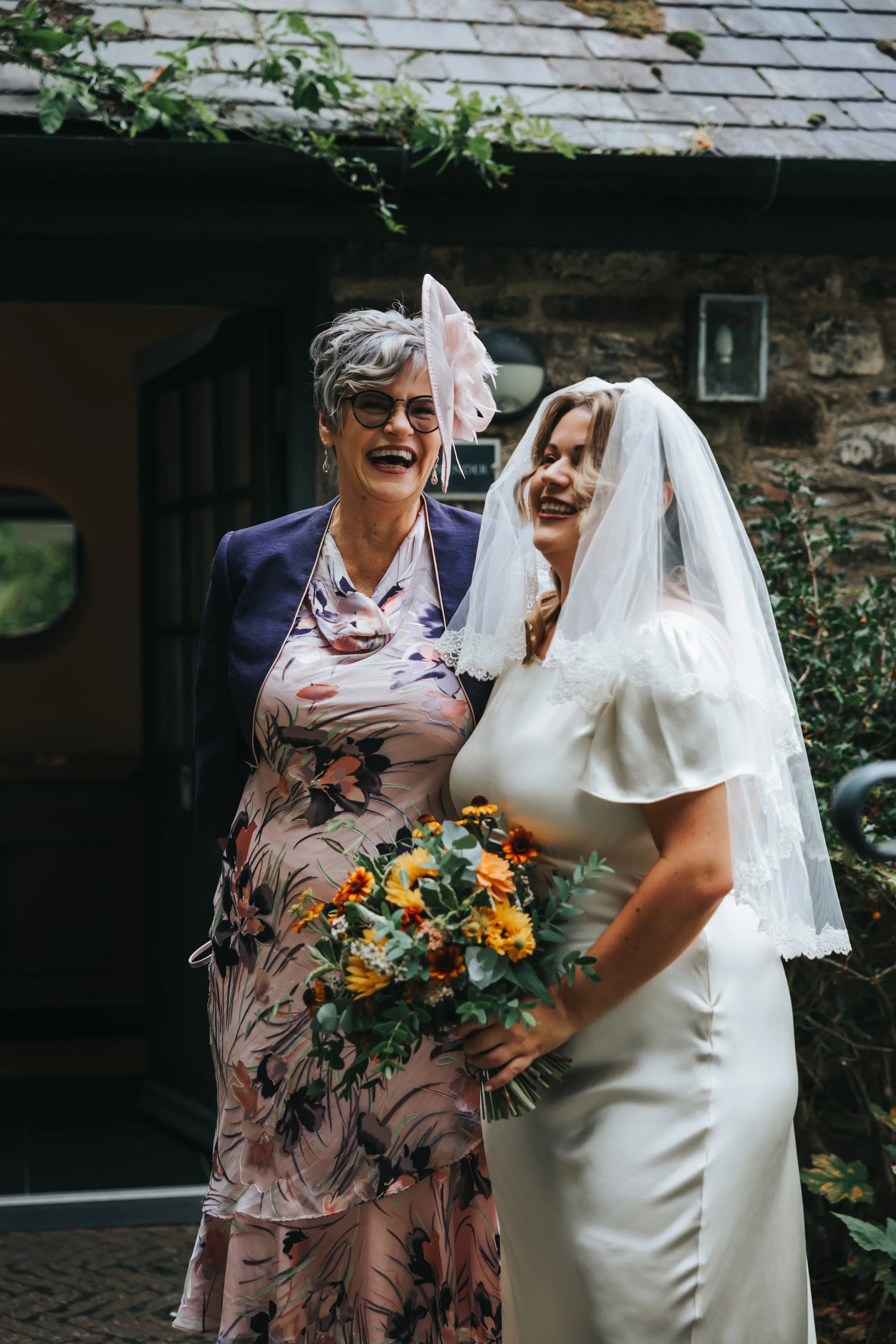 A bride and another woman laughing together outside a building, with the bride holding a bouquet of flowers.