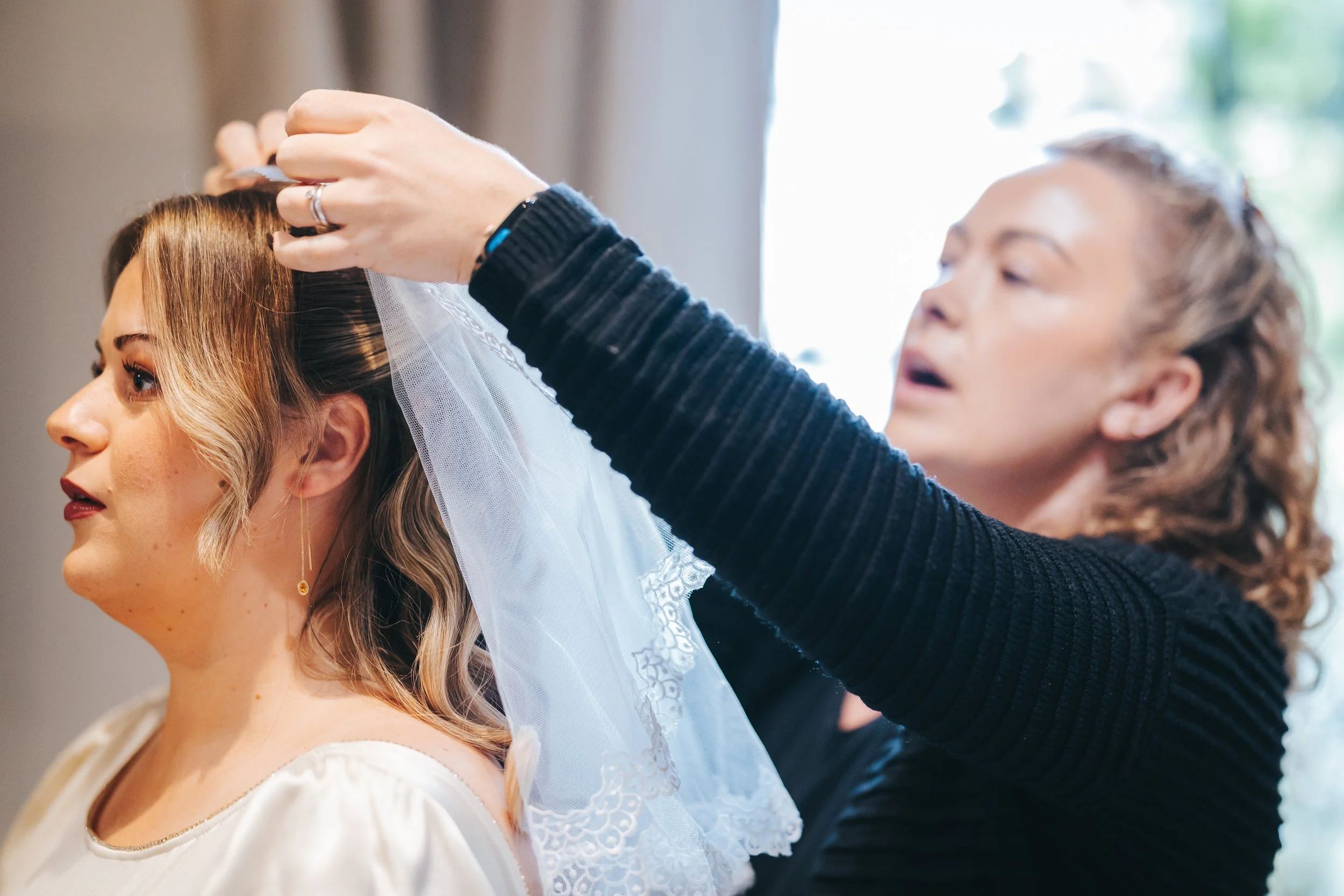 A woman helping another woman put on a wedding veil while getting ready.