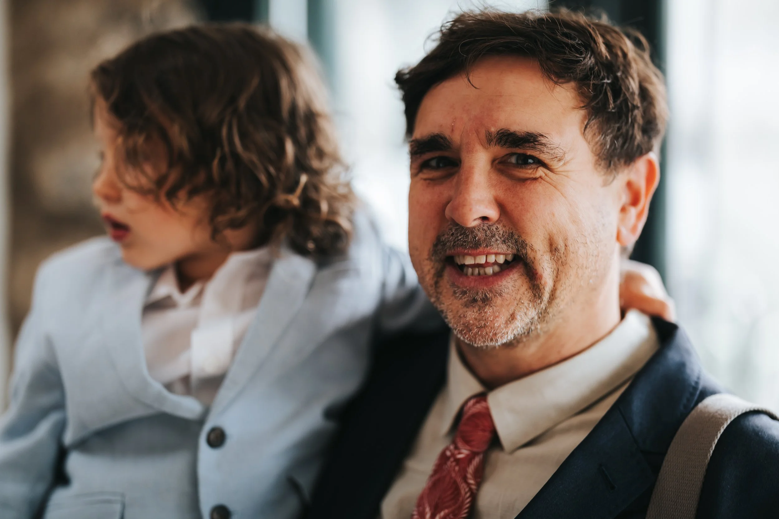 A man with short brown hair, facial hair, and wearing a suit and tie is smiling at the camera while carrying a child with curly brown hair and a gray jacket.