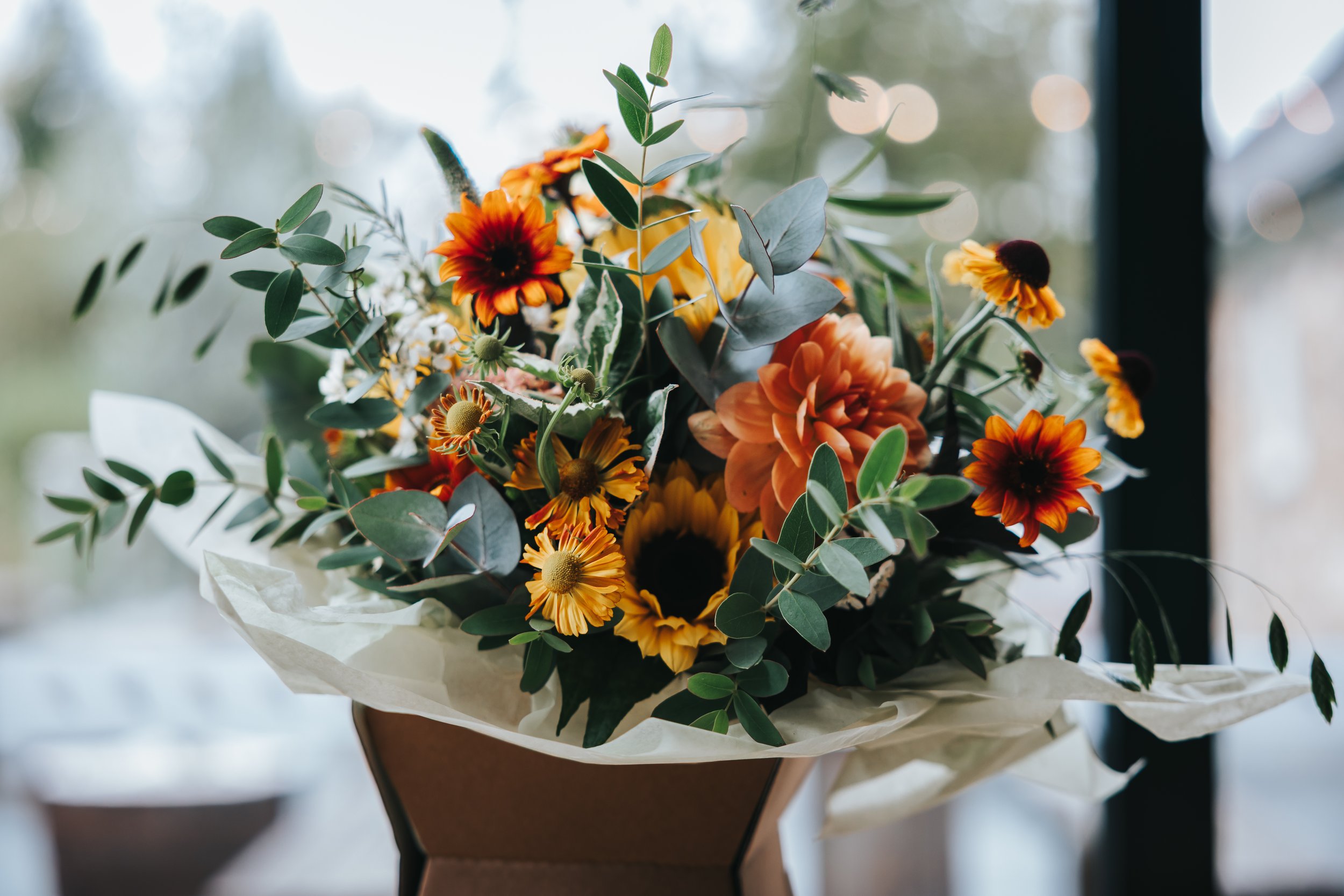 A bouquet of flowers with orange, yellow, and dark brown blooms, surrounded by green foliage, wrapped in white paper, placed in a brown box.
