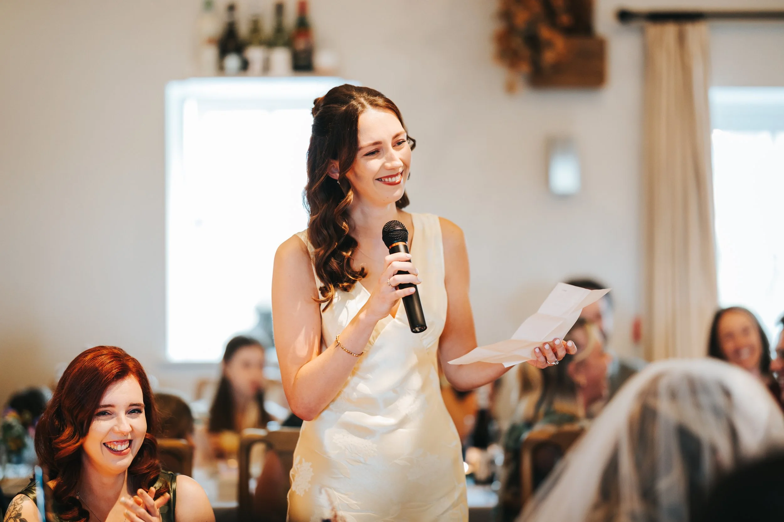 A woman giving a speech at a wedding reception, smiling and holding a microphone and notes, while seated guests join in the celebration.