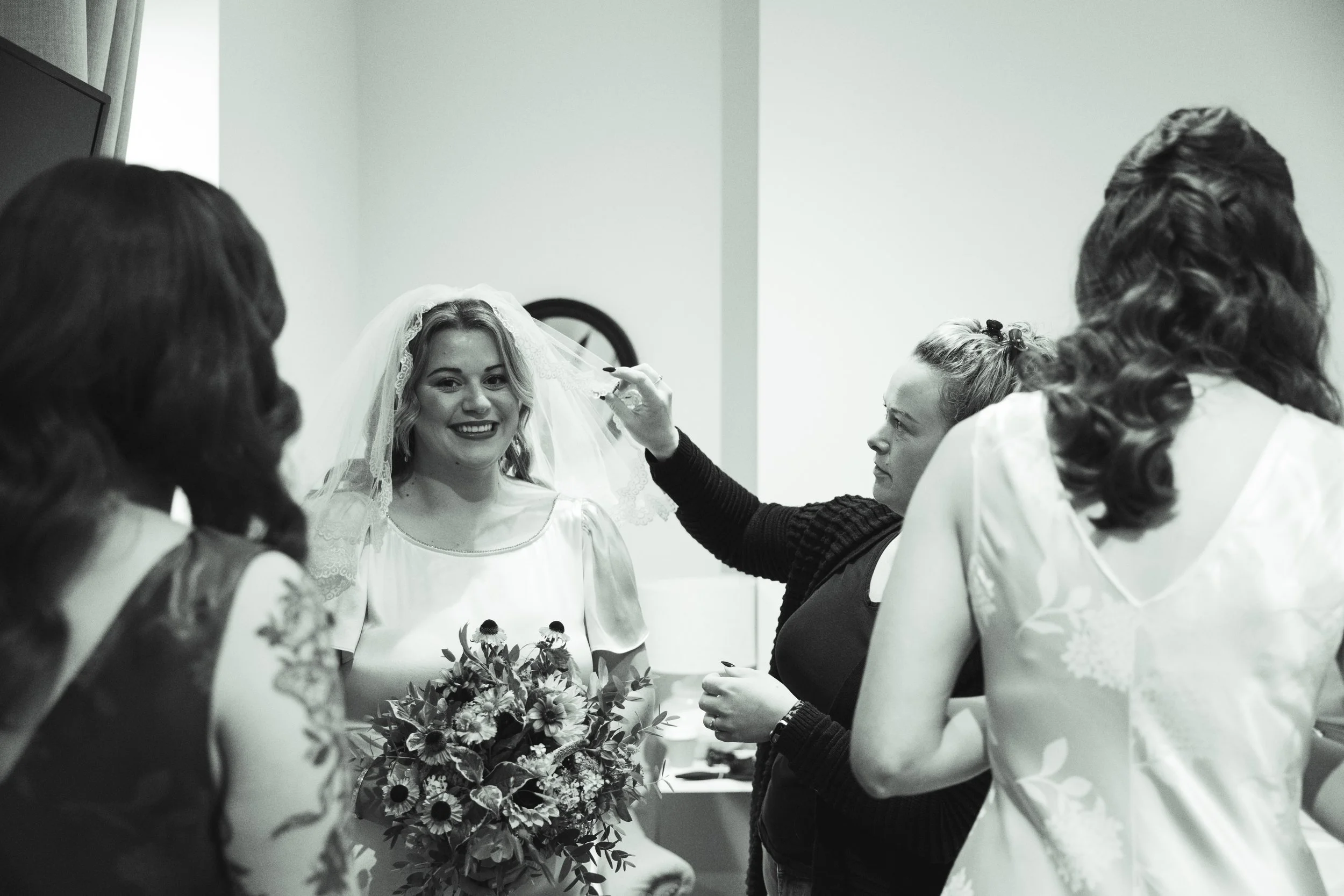 A bride is getting her veil adjusted by a woman, while three other women watch, in a dressing room.