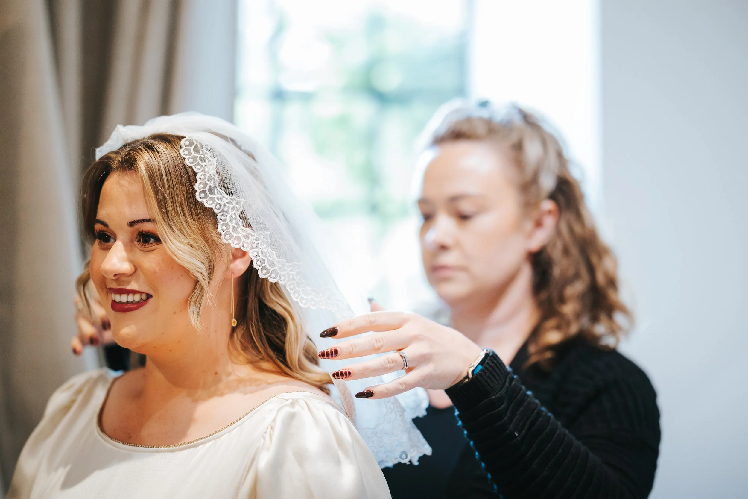 A bride with blonde hair smiling as she wears a lace veil, with a woman helping her prepare, possibly her stylist, in a softly lit room.