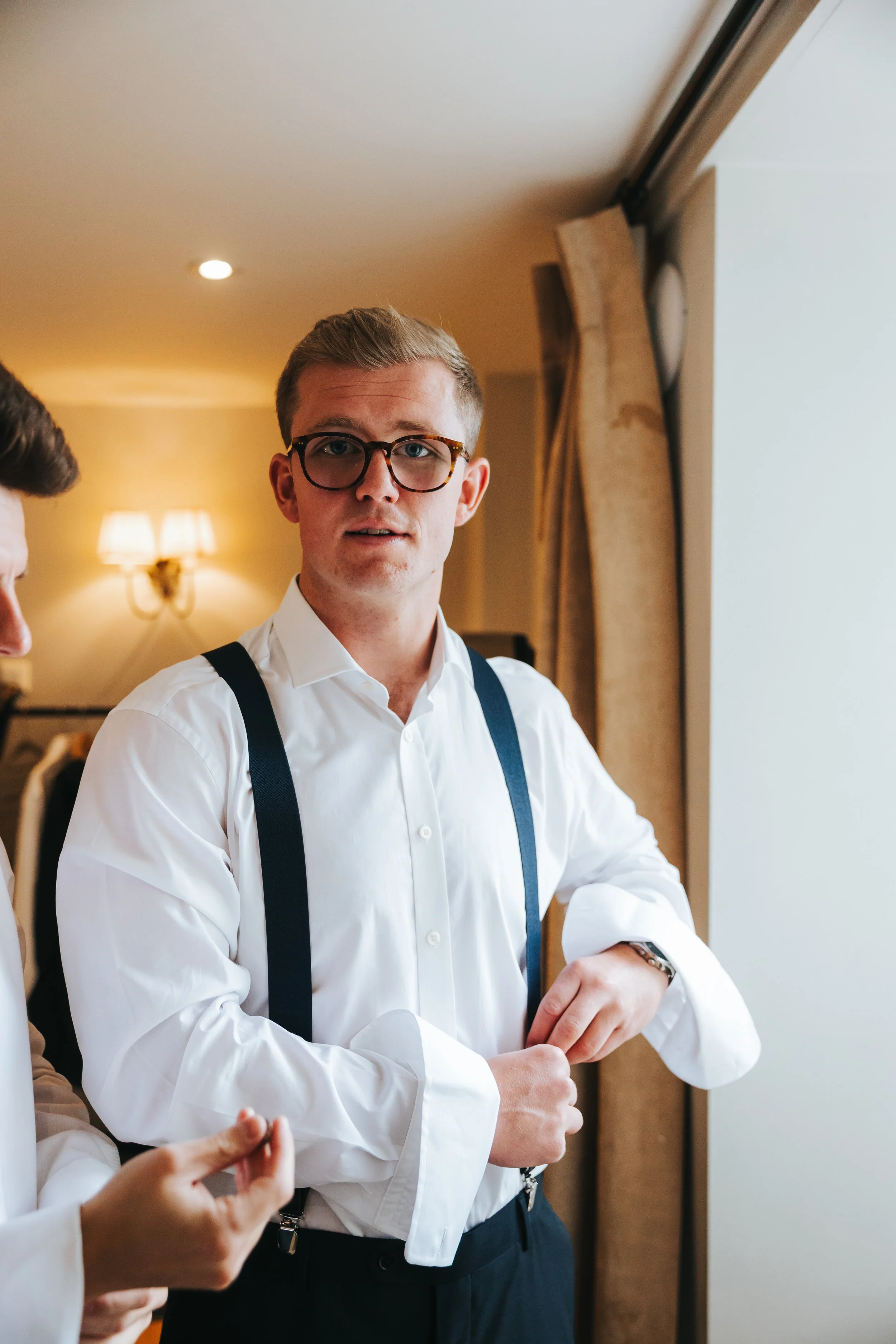 A young man with glasses and a white dress shirt with suspenders, standing indoors near a window, looking at the camera.