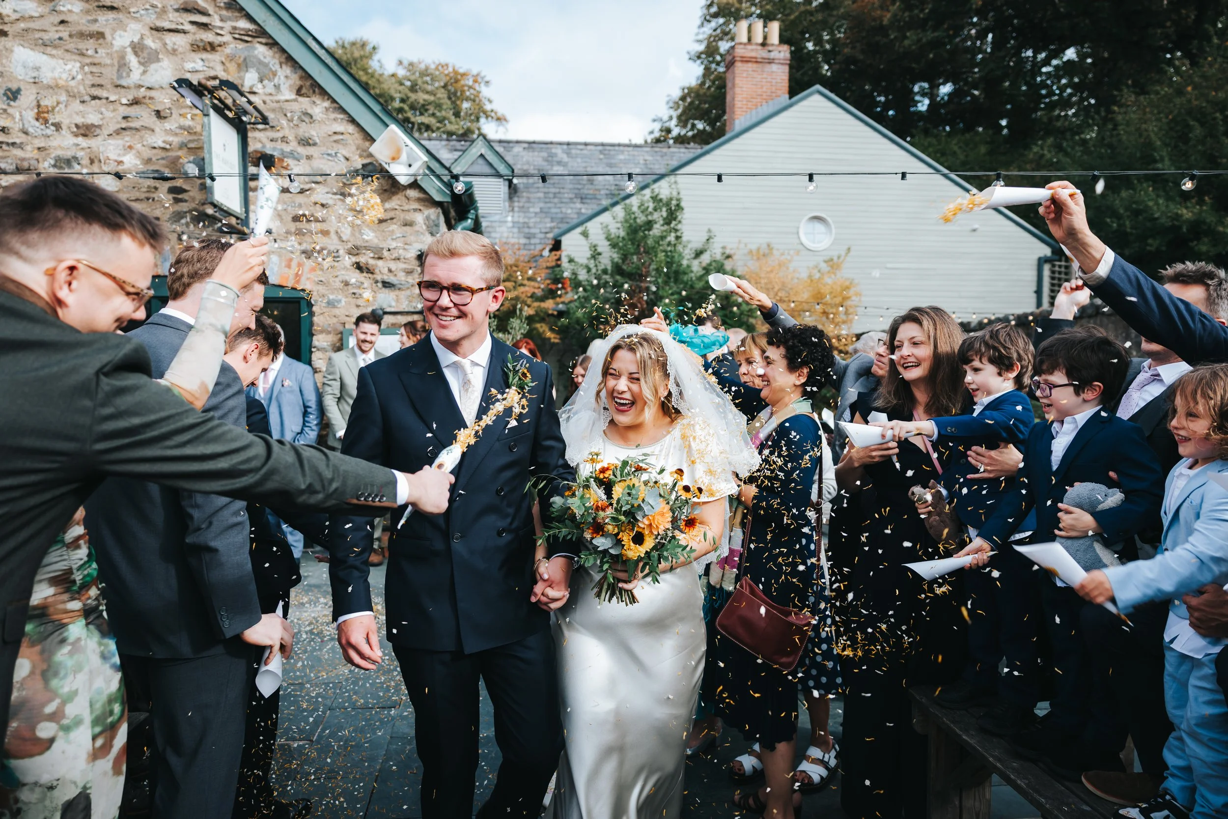Bride and groom walking hand in hand through a crowd celebrating with confetti and streamers at an outdoor wedding.