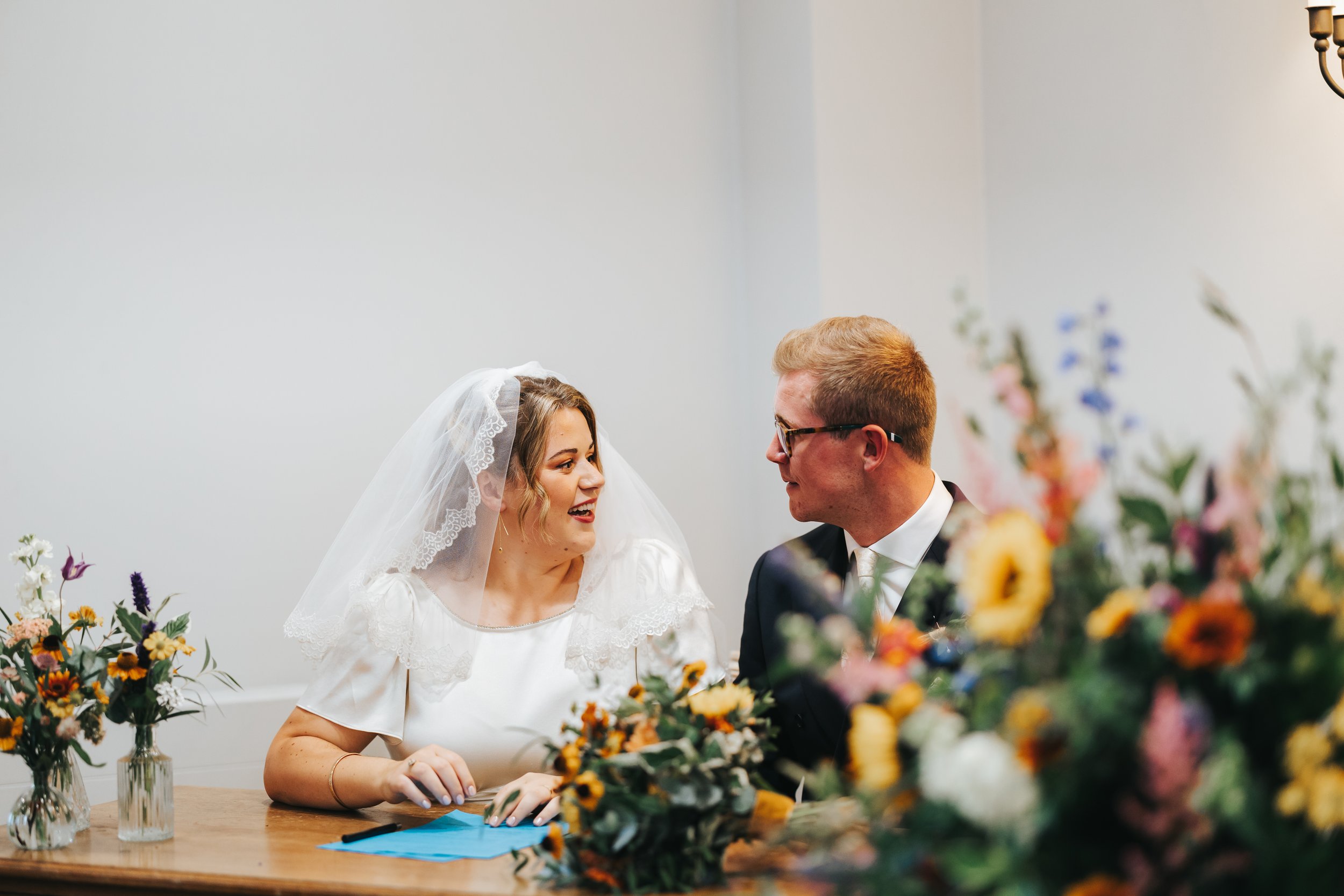 A bride and groom smiling and looking at each other during their wedding ceremony at a table decorated with colorful flowers and vases.