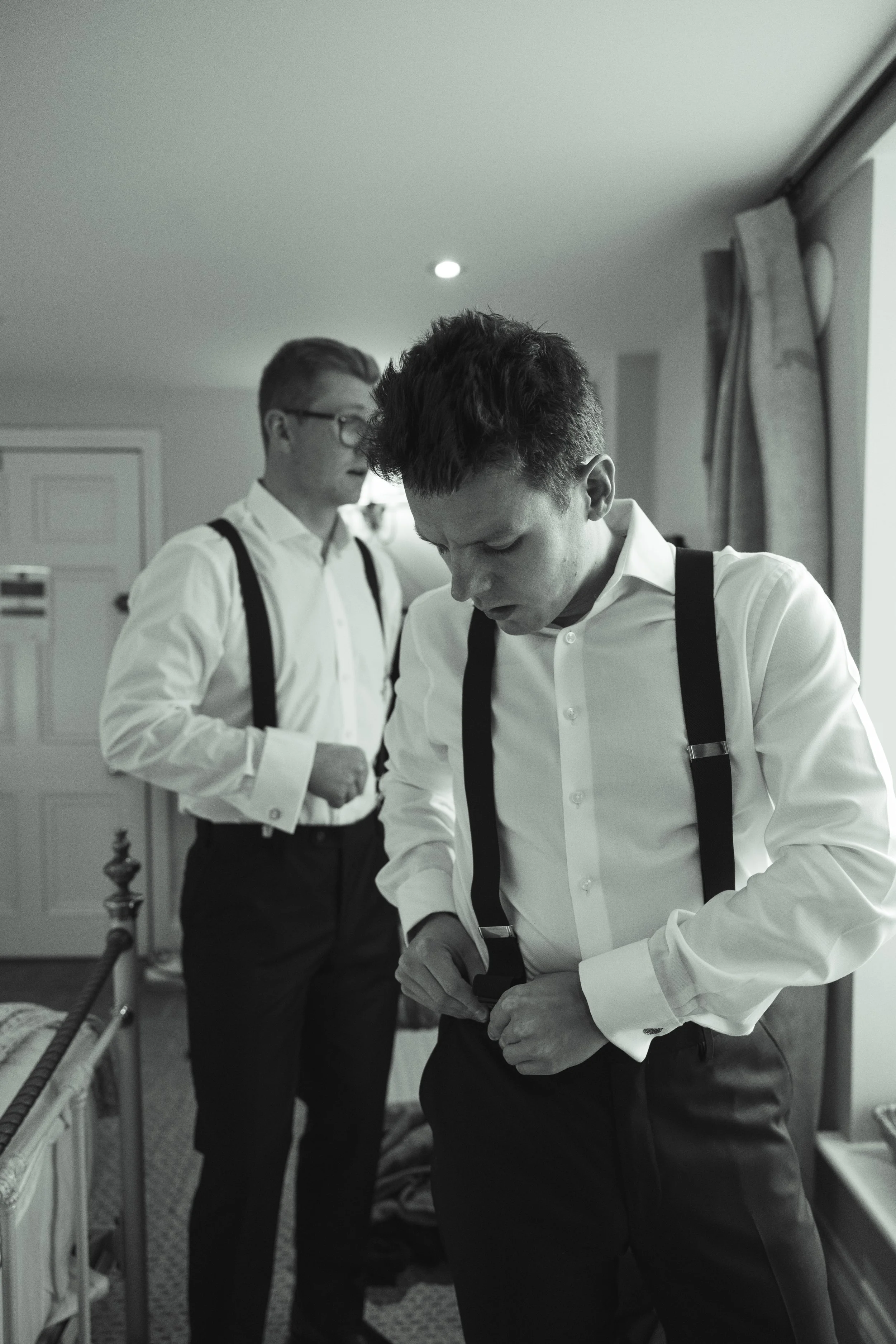 Two men in white dress shirts and black suspenders preparing for a formal event in a bedroom, with one adjusting his suspenders and the other looking down.
