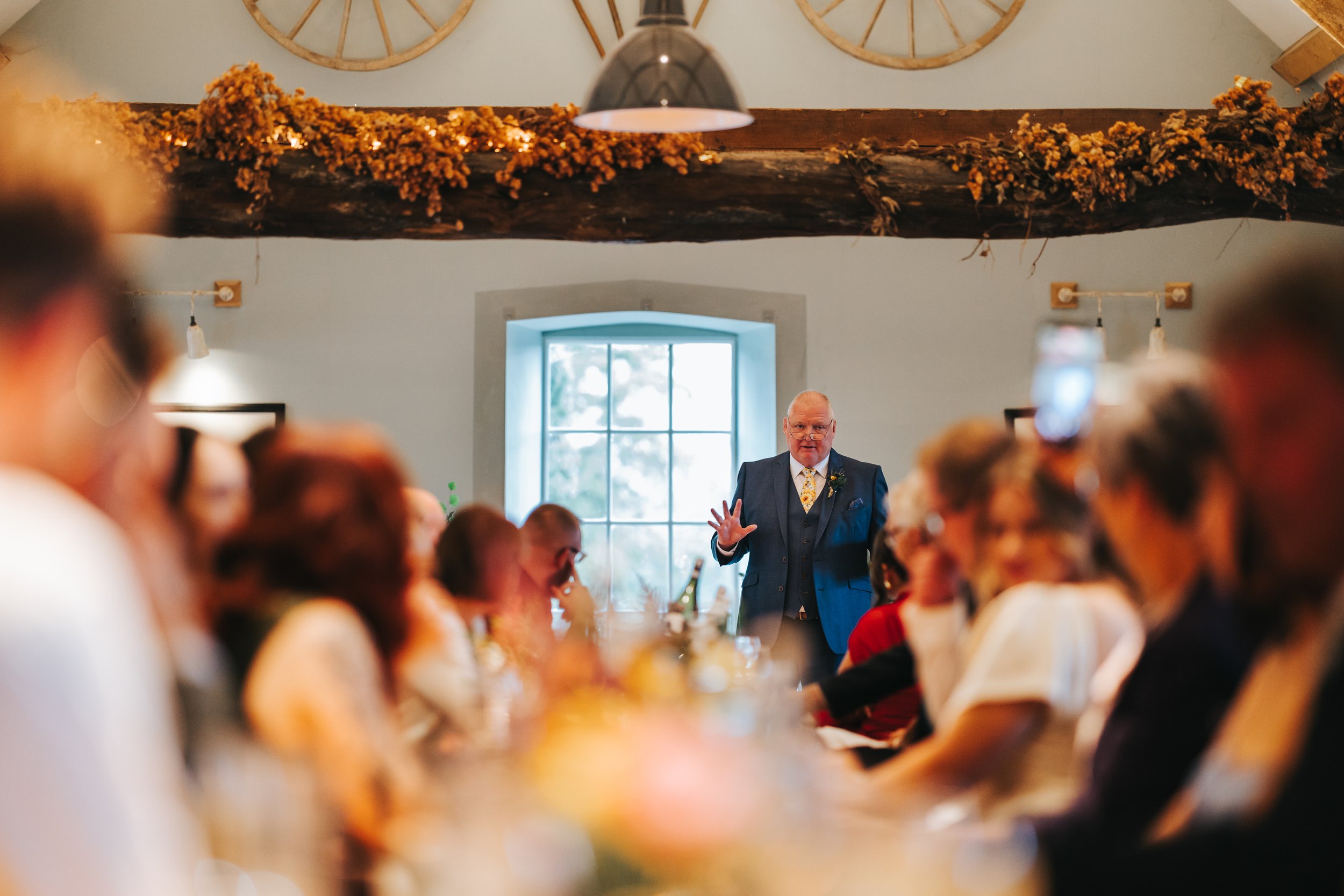 A man in a blue suit giving a speech at a gathering with guests seated at a long table, inside a rustic room with a window and wooden beam decorated with dried flowers.