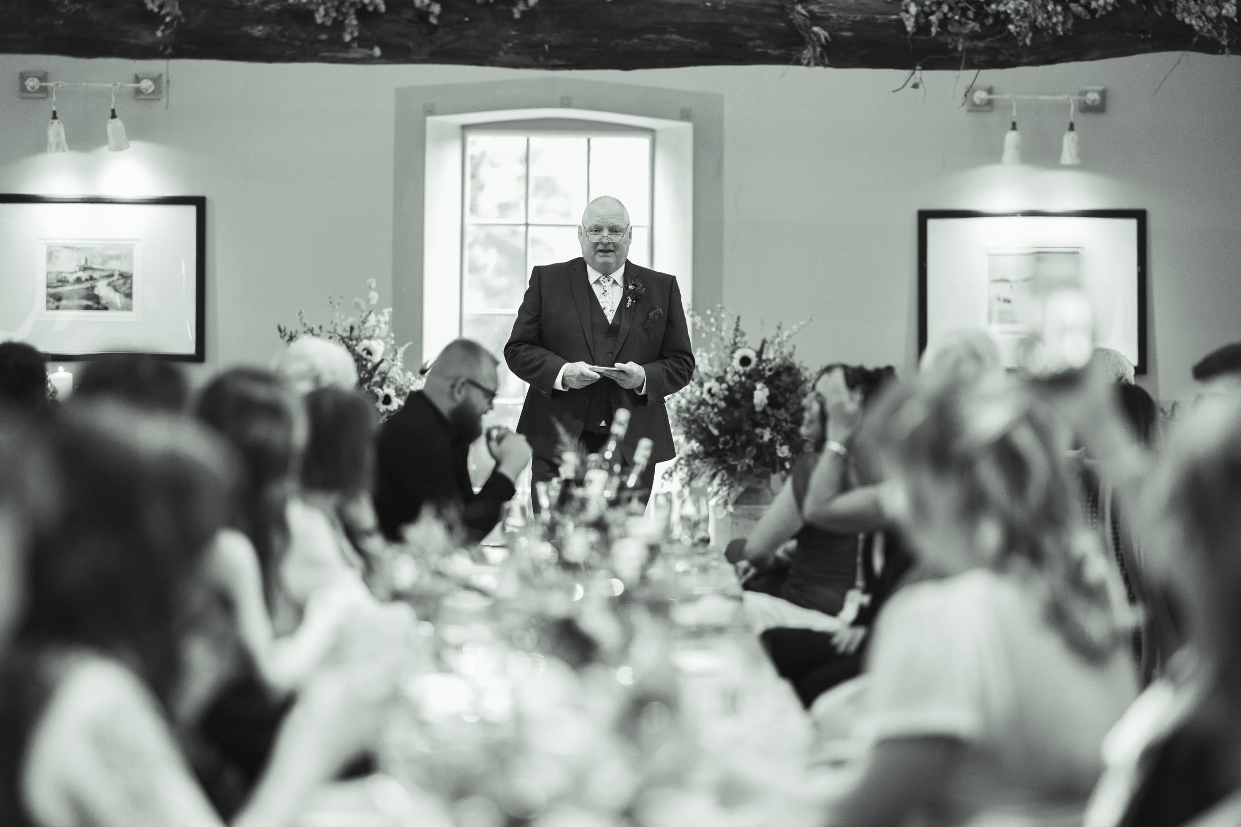 A man in a suit giving a speech at a formal gathering in a decorated room with floral arrangements and artwork, attended by seated guests.
