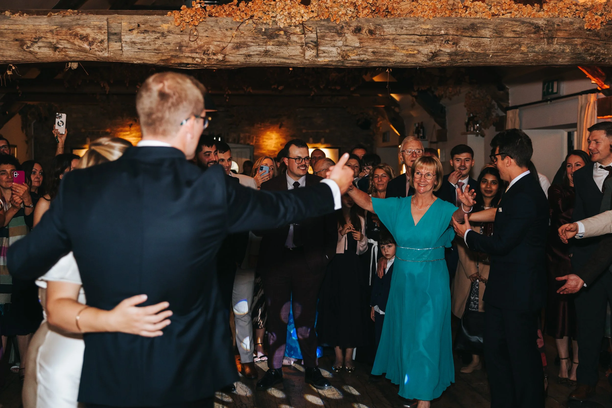 People dancing and celebrating at a wedding reception, with a woman in a teal dress in the center and guests taking photos.