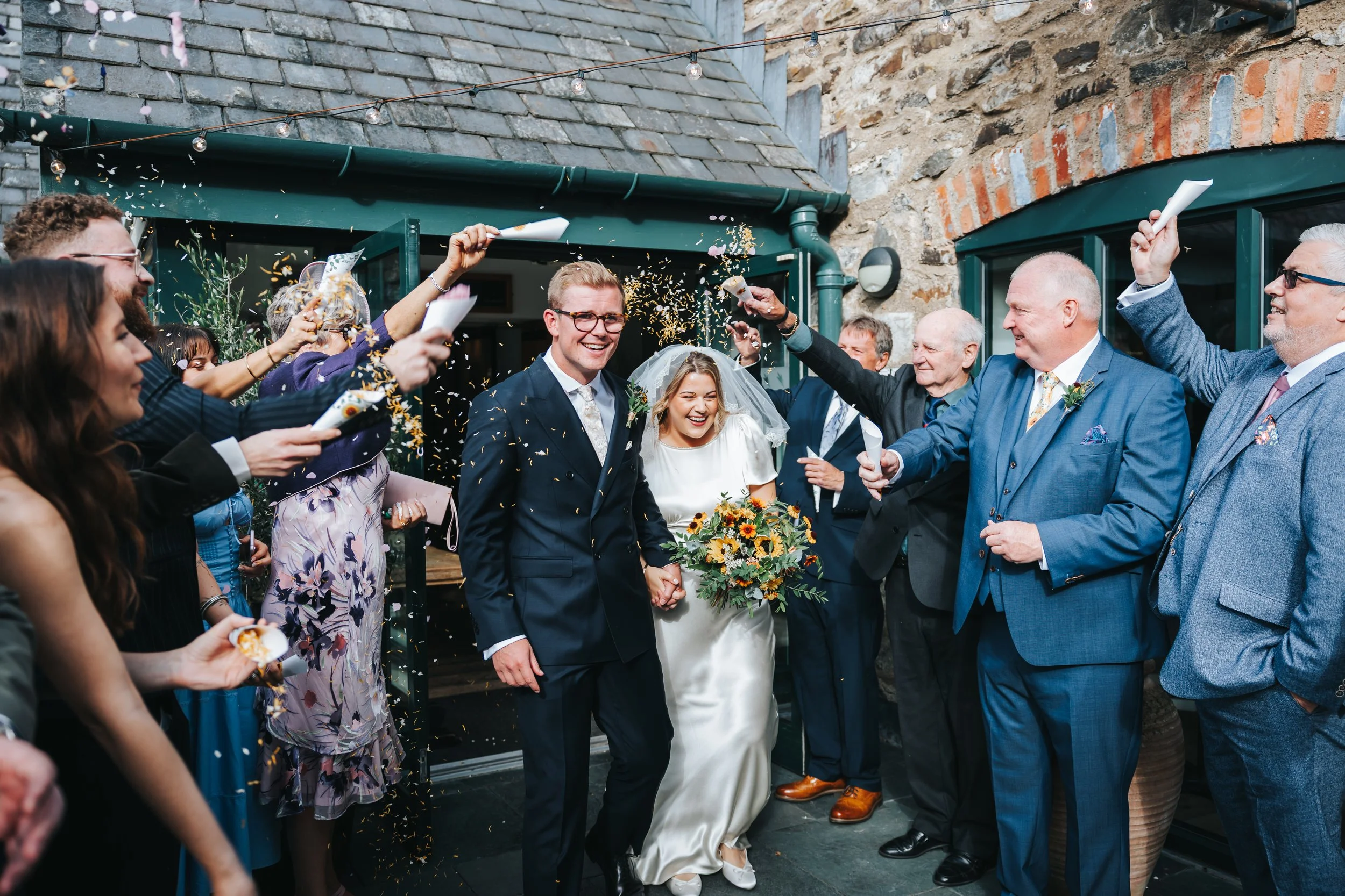 A newlywed couple exits a building while friends and family throw confetti and cheer. The groom wears a dark suit and glasses, holding the bride's hand. The bride wears a white dress, a veil, and holds a bouquet of sunflowers. The celebration takes p