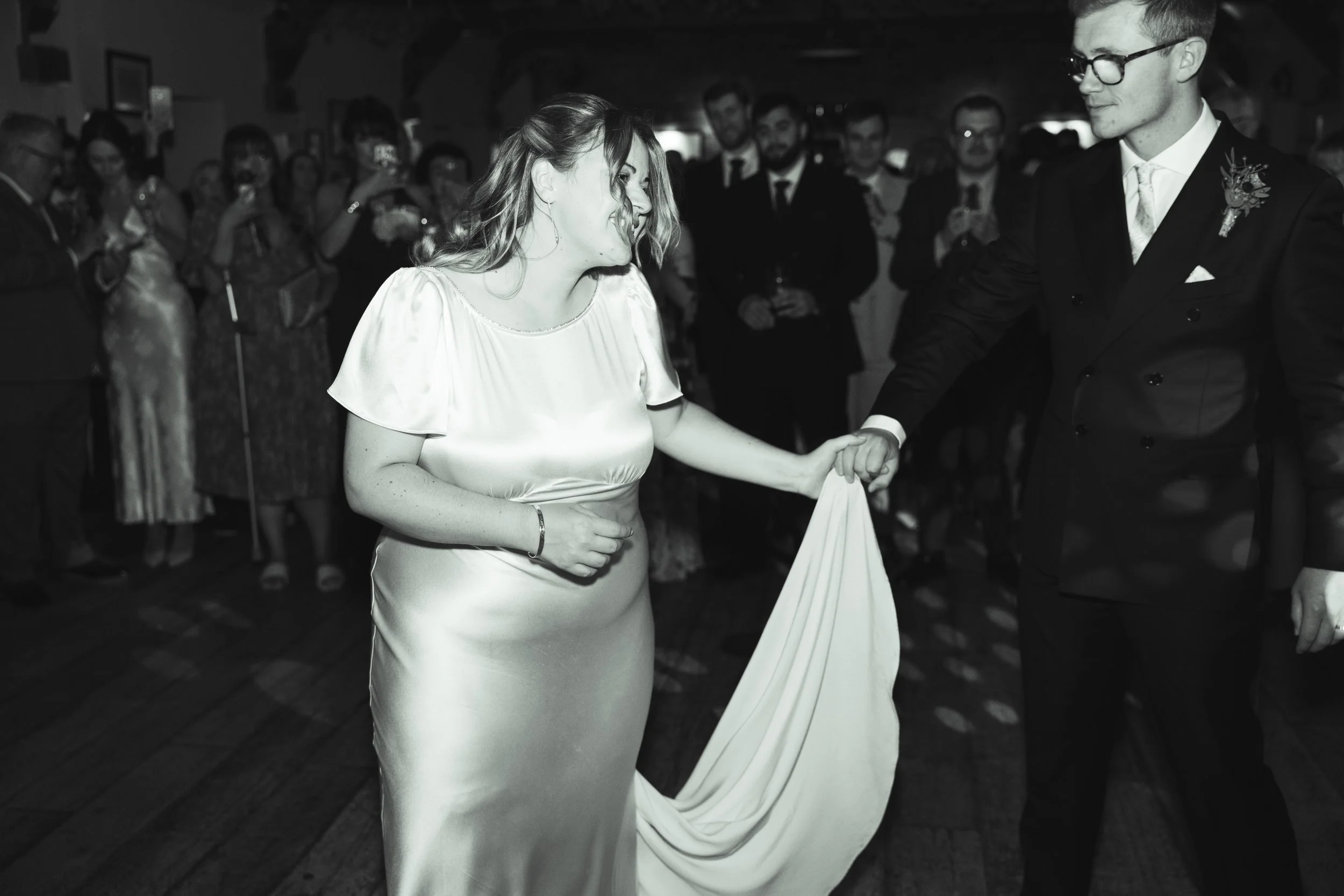 A black and white photo of a bride and groom dancing during their wedding reception. The bride is holding her wedding dress with one hand, and the groom is holding her hand. There are guests in the background watching and taking pictures.