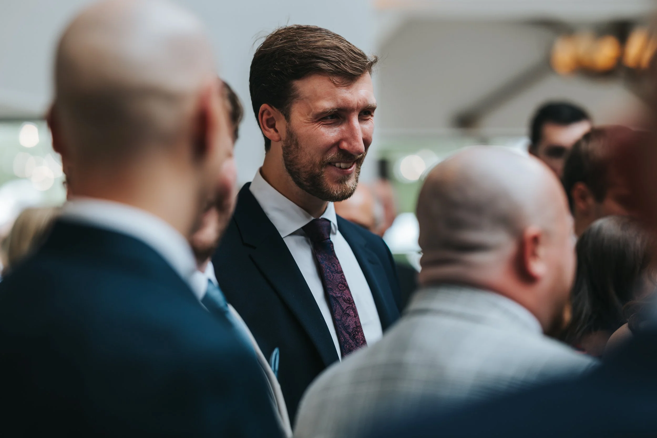 A man with a beard in formal attire, wearing a navy suit, while smiling and talking to people at a professional networking event or conference.