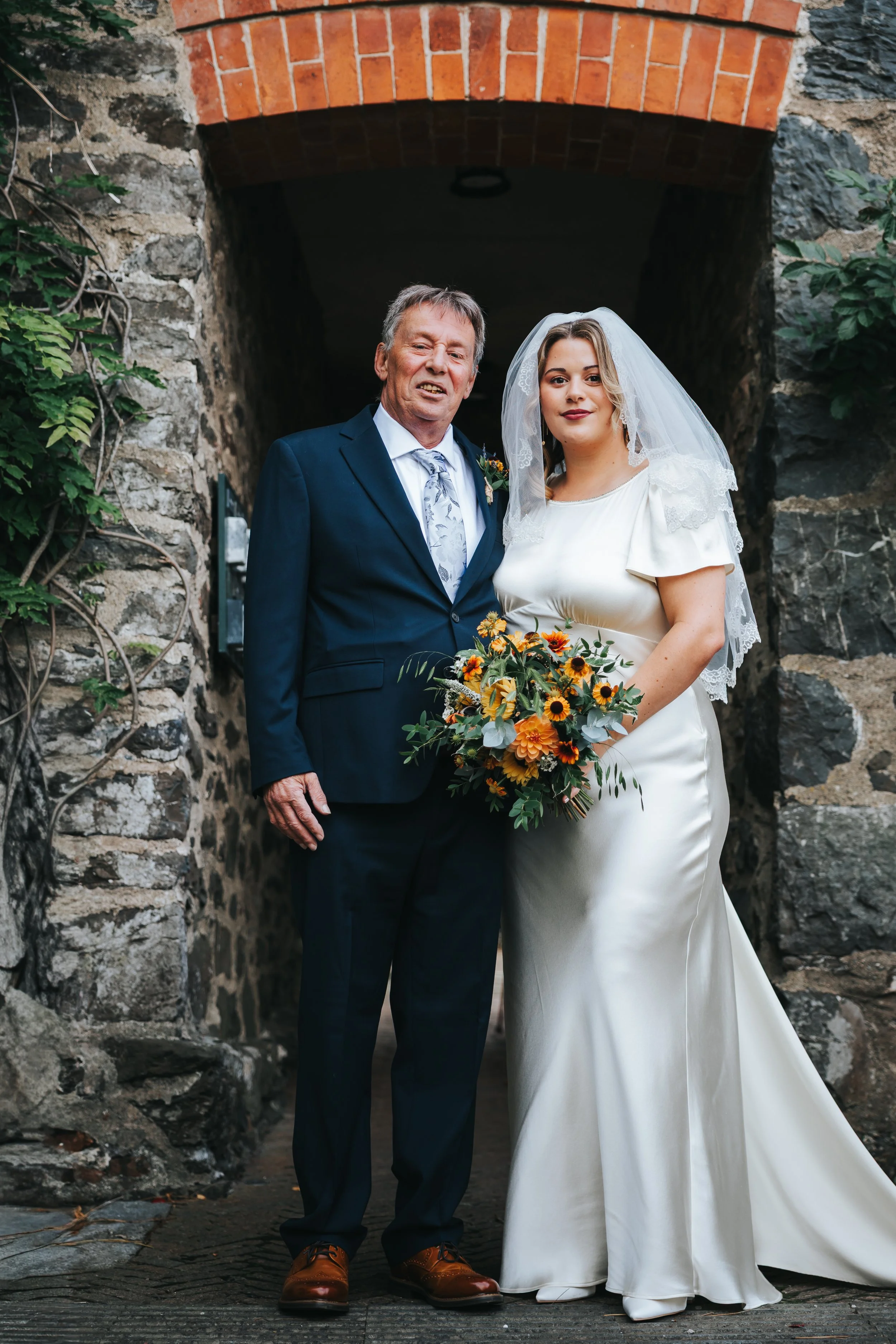 A bride in a white wedding gown holding a bouquet standing next to an older man in a blue suit, under a stone archway.