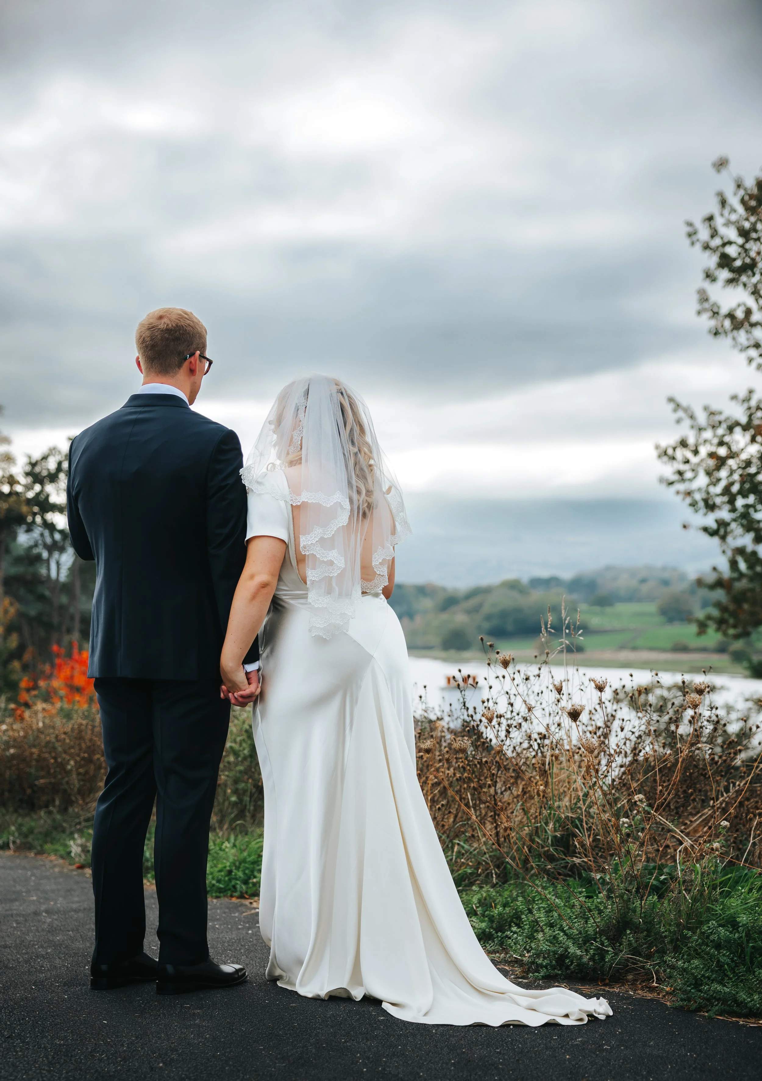 A bride and groom holding hands outdoors by a river under cloudy sky, with trees and hills in the background.