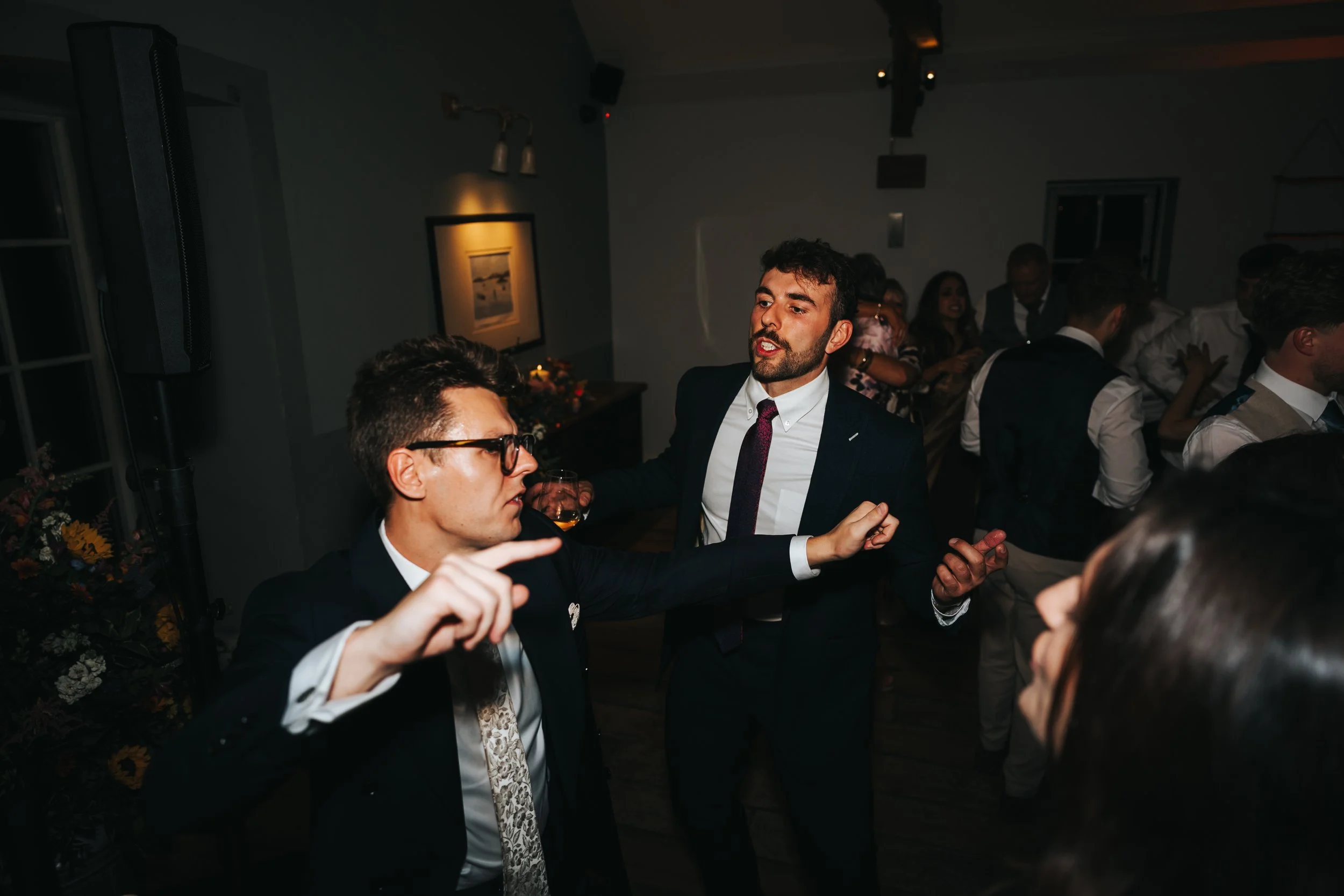 People dancing and socializing at a party, dressed in formal attire, in a dimly lit room with artwork on the walls and floral decorations.