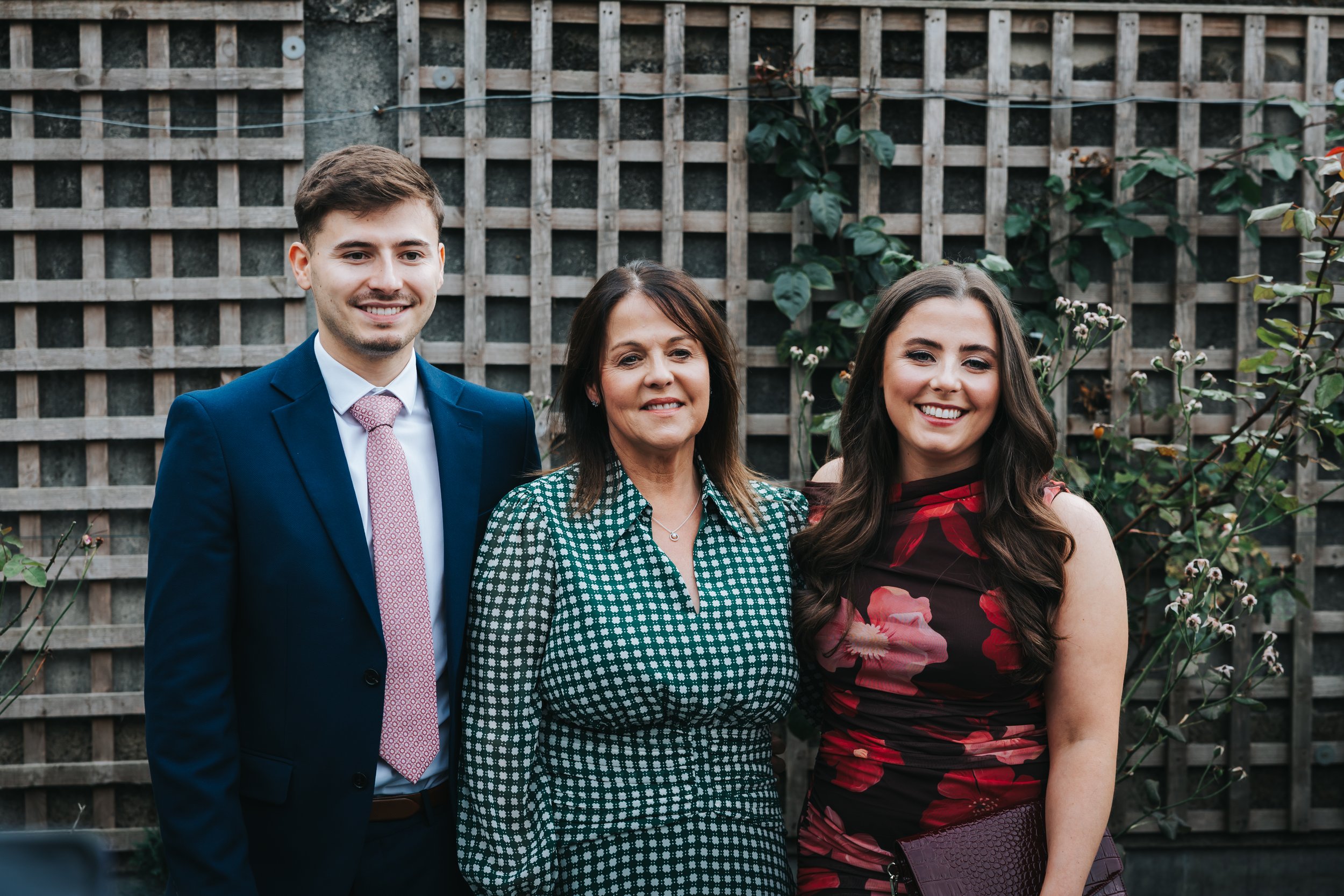 A family of three standing outdoors in front of a wooden lattice fence with green foliage. The man on the left is wearing a blue suit and a pink tie. The woman in the middle is wearing a green and white checked dress. The woman on the right is wearin