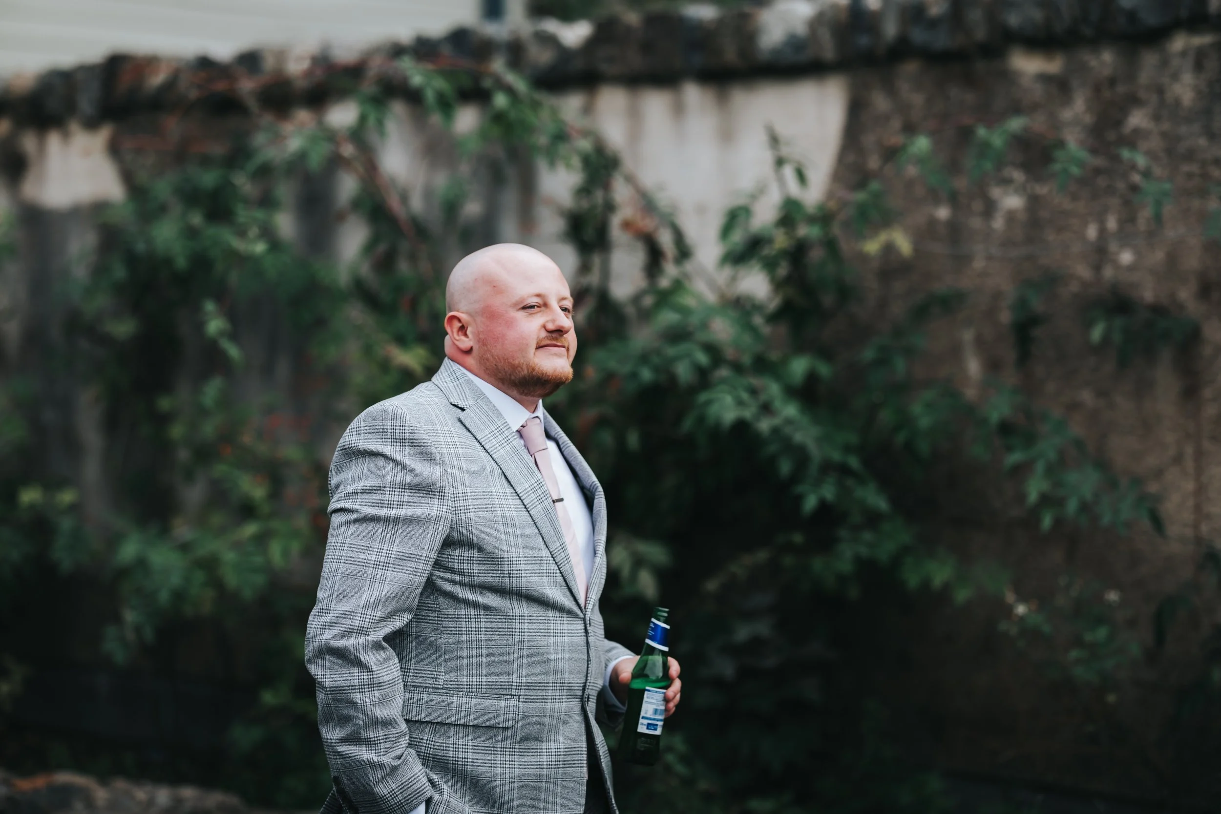 A bald man with a beard wearing a light gray plaid suit and a pink tie, standing outdoors. He is holding a beer bottle and looking to the side with a confident expression. There is a background of a brick wall and green foliage.