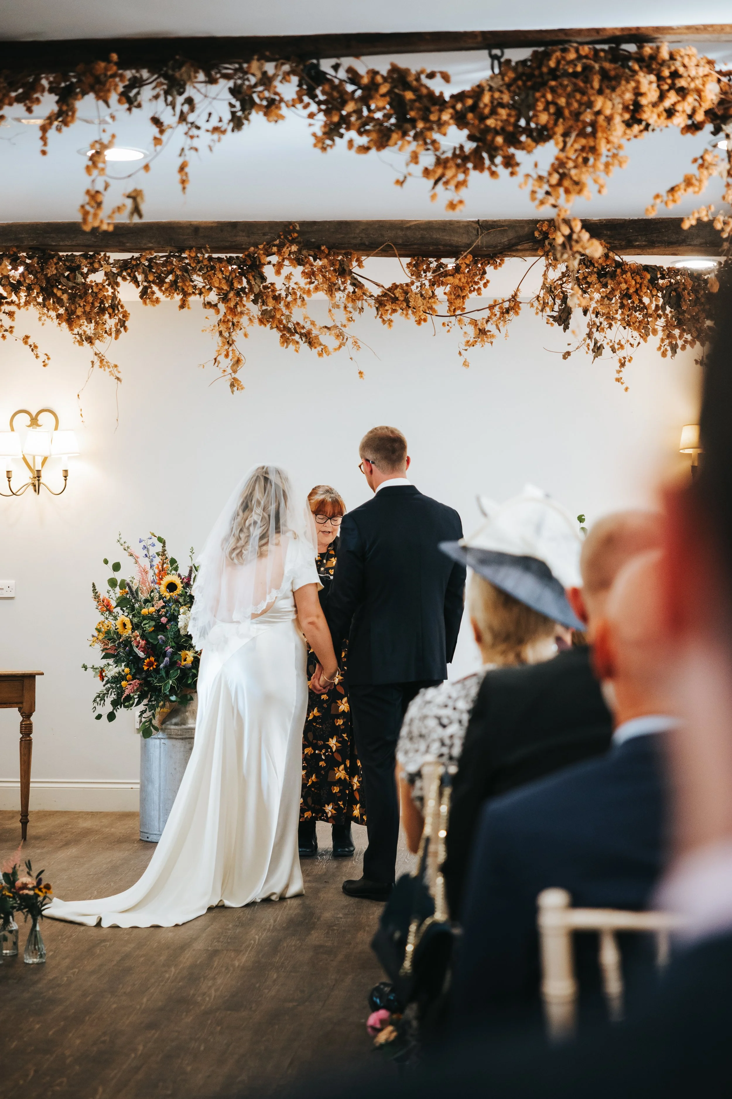 A wedding ceremony with a bride and groom standing before an officiant, holding hands, in a decorated room with flower arrangements and dried floral garlands overhead.