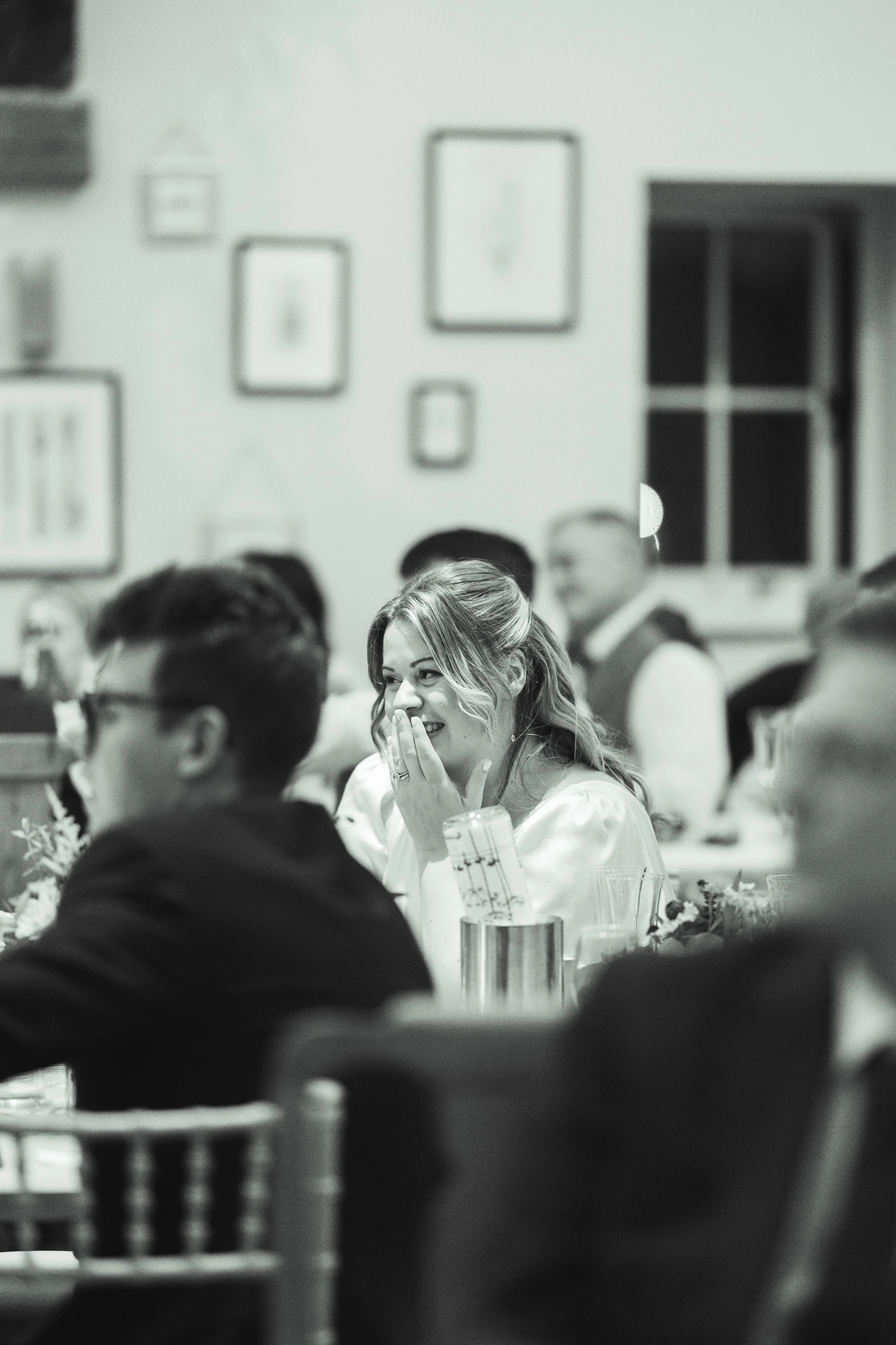 A woman with wavy hair sitting at a table, smiling and covering her mouth with her hand, in a gathering or celebration setting.