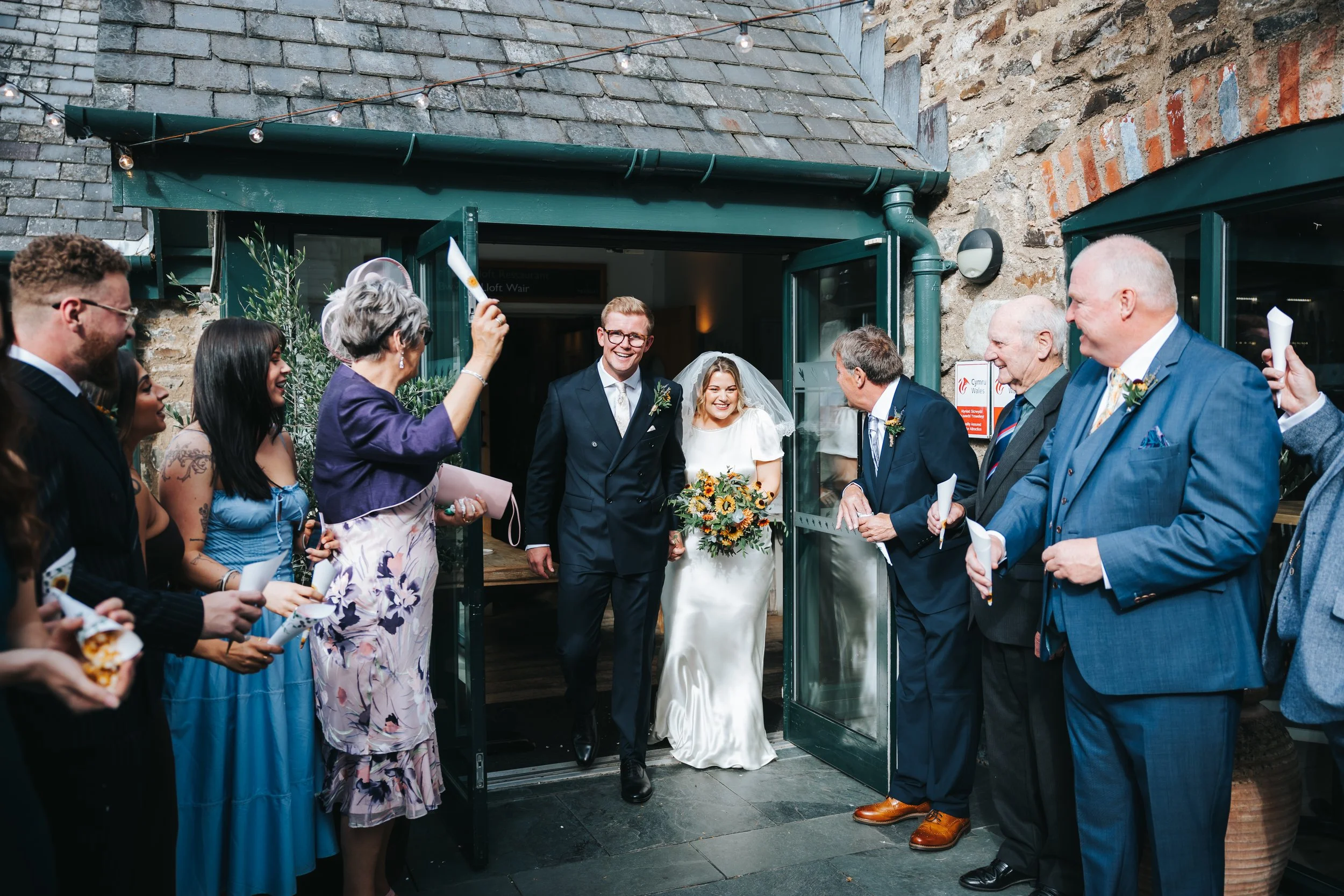 A wedding celebration with the bride and groom exiting a venue, surrounded by friends and family who are smiling and holding papers, with a woman in a purple dress waving a paper. The scene takes place outside a rustic stone building with open green 