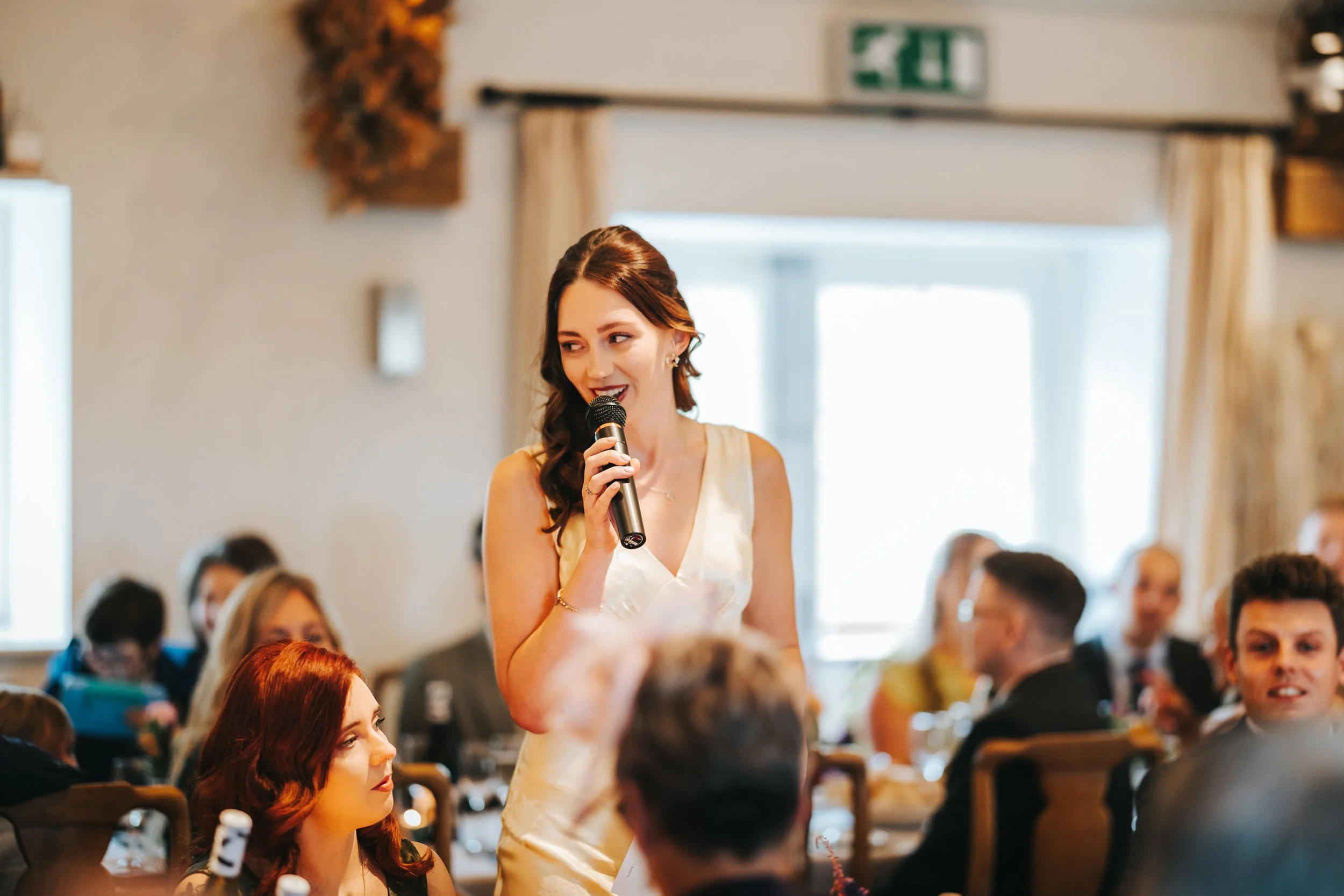 Woman in champagne dress giving speech with microphone at wedding reception, group of guests seated at tables in background.