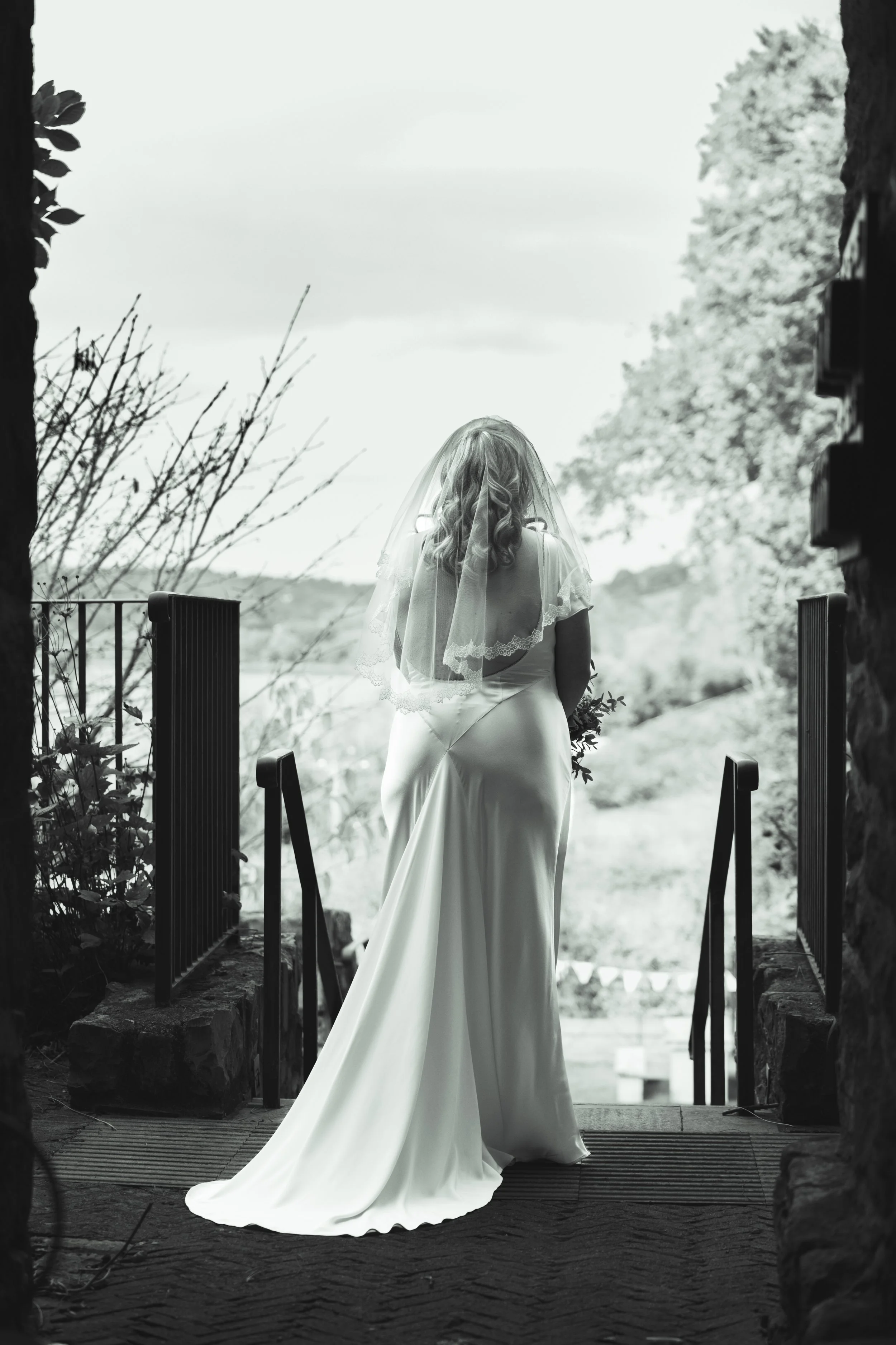 A woman in a white wedding dress and veil stands at an entrance, looking out over a landscape with trees and a body of water in the distance.