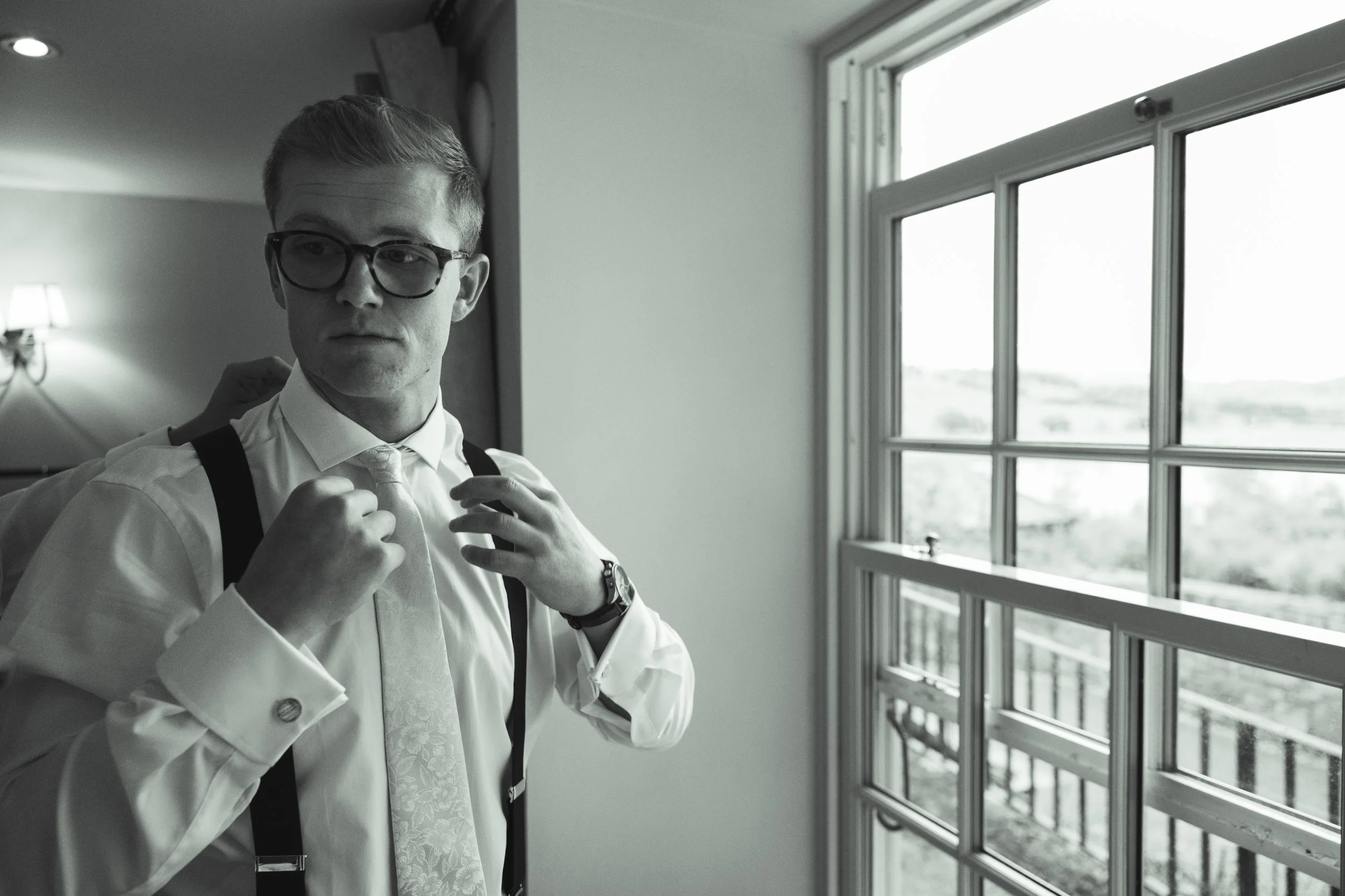 A man with glasses, wearing a white shirt, tie, and suspenders, adjusting his tie while looking out a window, with an uncluttered indoor setting and natural light coming through the window.